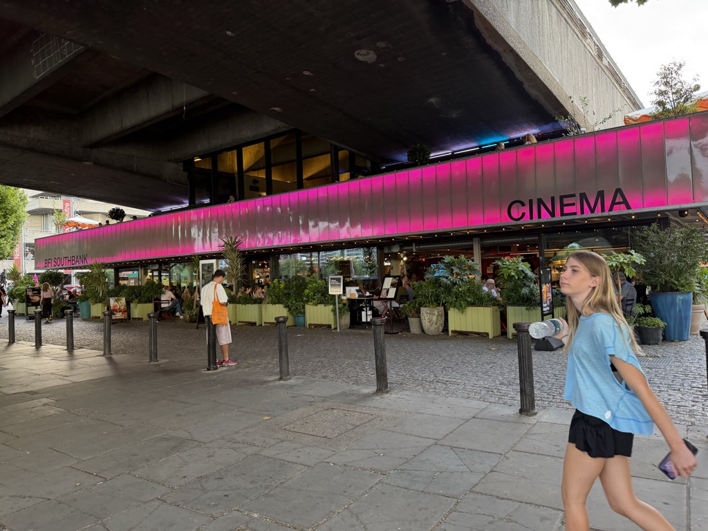 The BFI Southbank cinema in London tucked beneath the concrete underside of Waterloo Bridge, its frontage lit by a bright magenta illuminated panel with black lettering reading 'Cinema' and 'BFI Southbank'. Potted plants and green planters edge an outdoor seating area where people sit at tables, and pedestrians walk across the paved terrace in the foreground