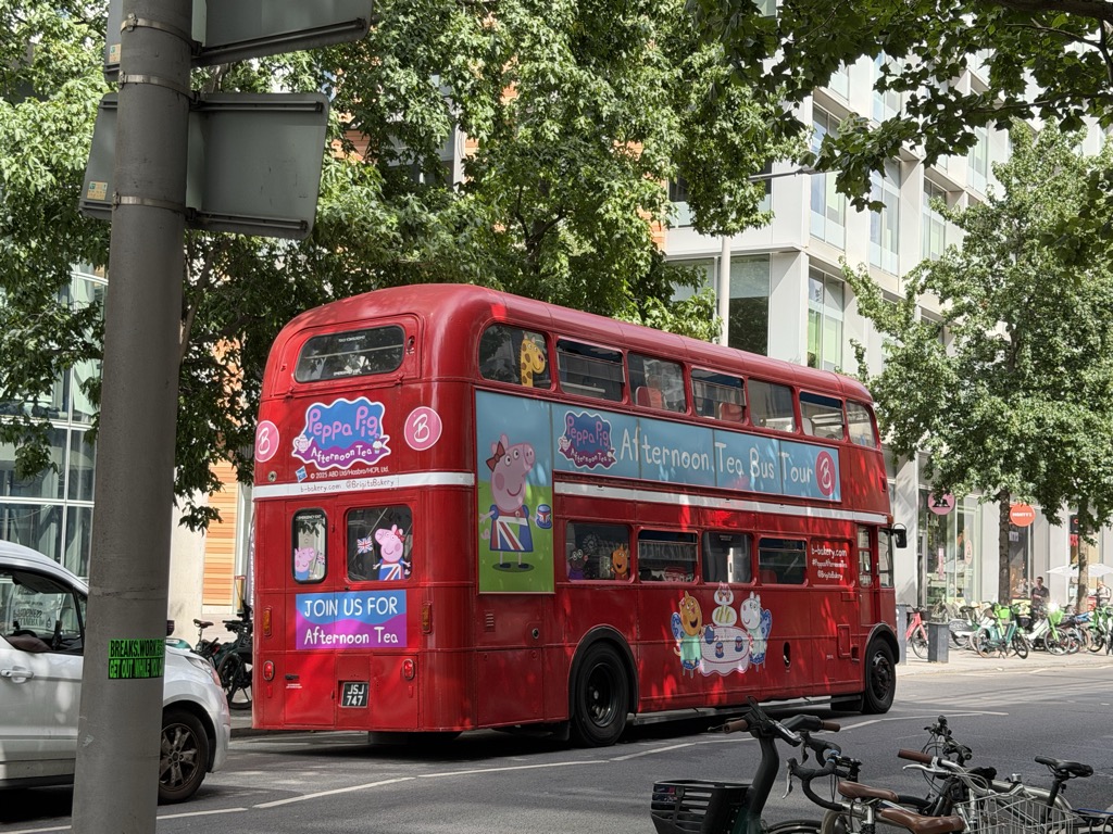 A red London Routemaster double-decker bus wrapped in Peppa Pig Afternoon Tea Bus Tour advertising, parked on a tree-lined street. The rear panel reads 'Join us for Afternoon Tea' and features Peppa Pig characters at a tea party, with registration plate JSJ 747. Parked bicycles, a rental bike rack, and modern glass apartment buildings line the background