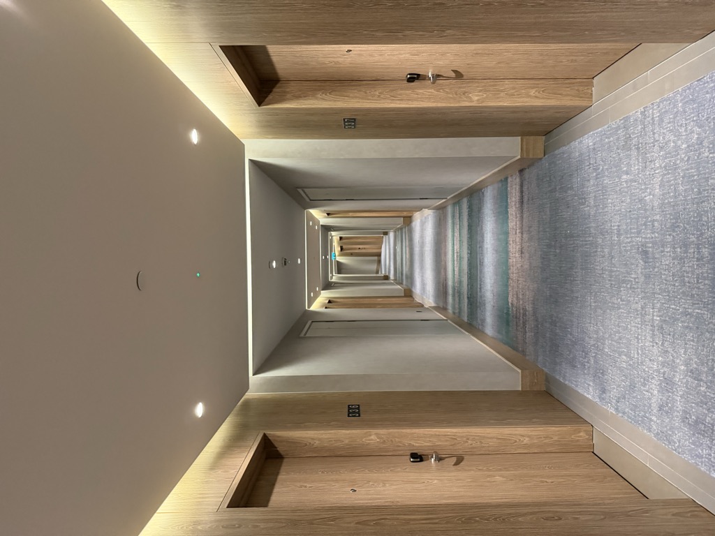 Empty hotel guest room corridor with pale oak door surrounds and recessed ceiling lighting, receding in one-point perspective toward a distant green exit sign. Room numbers are mounted beside each door and a grey and teal patterned carpet runner runs down the center of the tiled floor