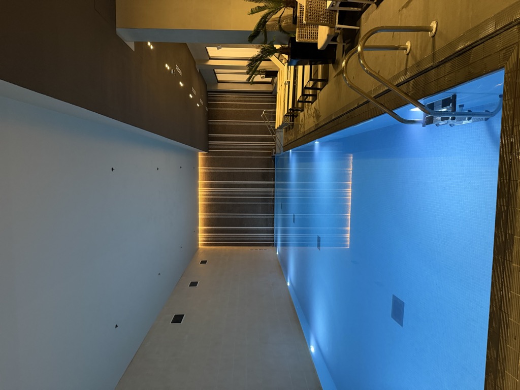 Indoor hotel swimming pool on a basement level at the Hilton London Bankside, lit pale blue from underwater lights, with a stainless steel handrail and ladder at the near corner. Wicker loungers and chairs with cream cushions line the right-hand side beneath potted palms, and a backlit vertical slatted wall glows warm at the far end, reflecting on the still water