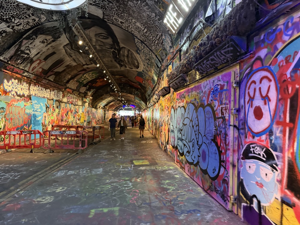 Inside the Leake Street Arches graffiti tunnel near Waterloo Station, a long curved railway underpass covered floor to ceiling in layered spray-painted murals and tags in bright pinks, blues, and yellows. A large monochrome portrait of a face is painted across the ceiling vault, cartoon characters cover the right-hand wall, and several people walk toward the daylight at the far end past red plastic barriers