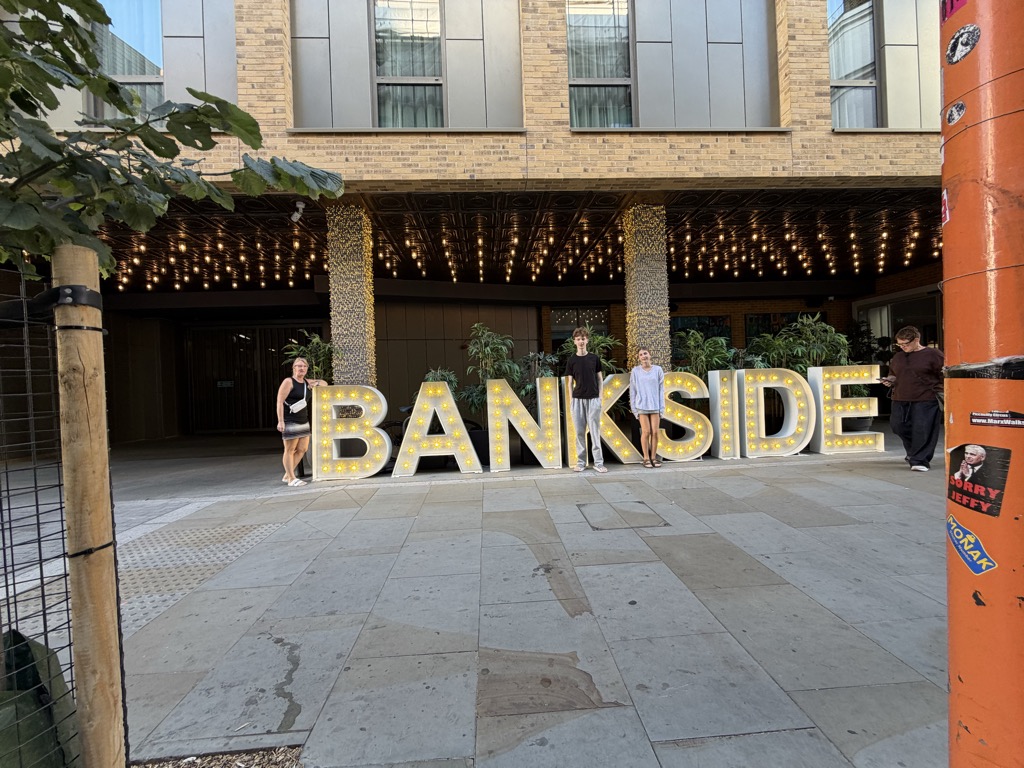 Large freestanding letters spelling BANKSIDE, each studded with round bulbs, set up on a paved plaza outside a modern brick and glass building whose entrance canopy is hung with strings of warm fairy lights. Three people pose among the letters and a passerby walks by on the right beside an orange bollard covered in stickers