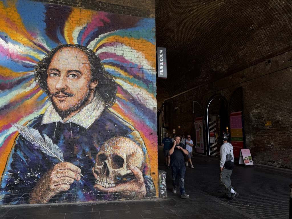 A large painted mural of William Shakespeare on a brick railway arch wall in Bankside, showing him holding a quill in one hand and a skull in the other against a starburst of orange, pink, blue, and purple rays. Pedestrians walk through the dim brick underpass to the right past a wagamama sign and illuminated shopfronts