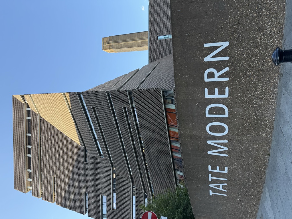 The Tate Modern seen from street level, with the angular brown brick Blavatnik Building rising in stepped, twisting tiers against a clear blue sky and the original power station's tall chimney visible behind it to the right. In the foreground a curved concrete retaining wall carries the gallery name 'TATE MODERN' in large white letters