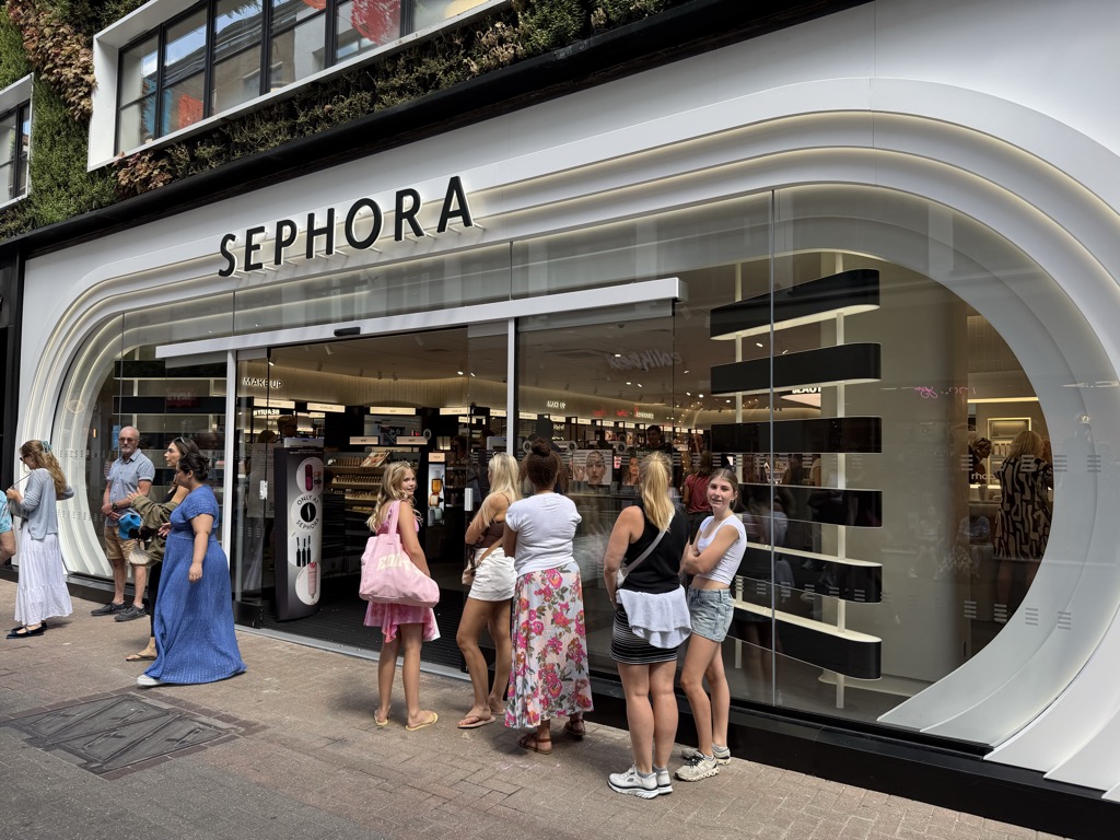 The Sephora storefront on Carnaby Street, a curved white facade with a large arched glass window and black lettering spelling SEPHORA, with a living green wall on the floor above. A queue of shoppers waits on the pavement outside the open entrance, and cosmetics displays and browsing customers are visible through the glass