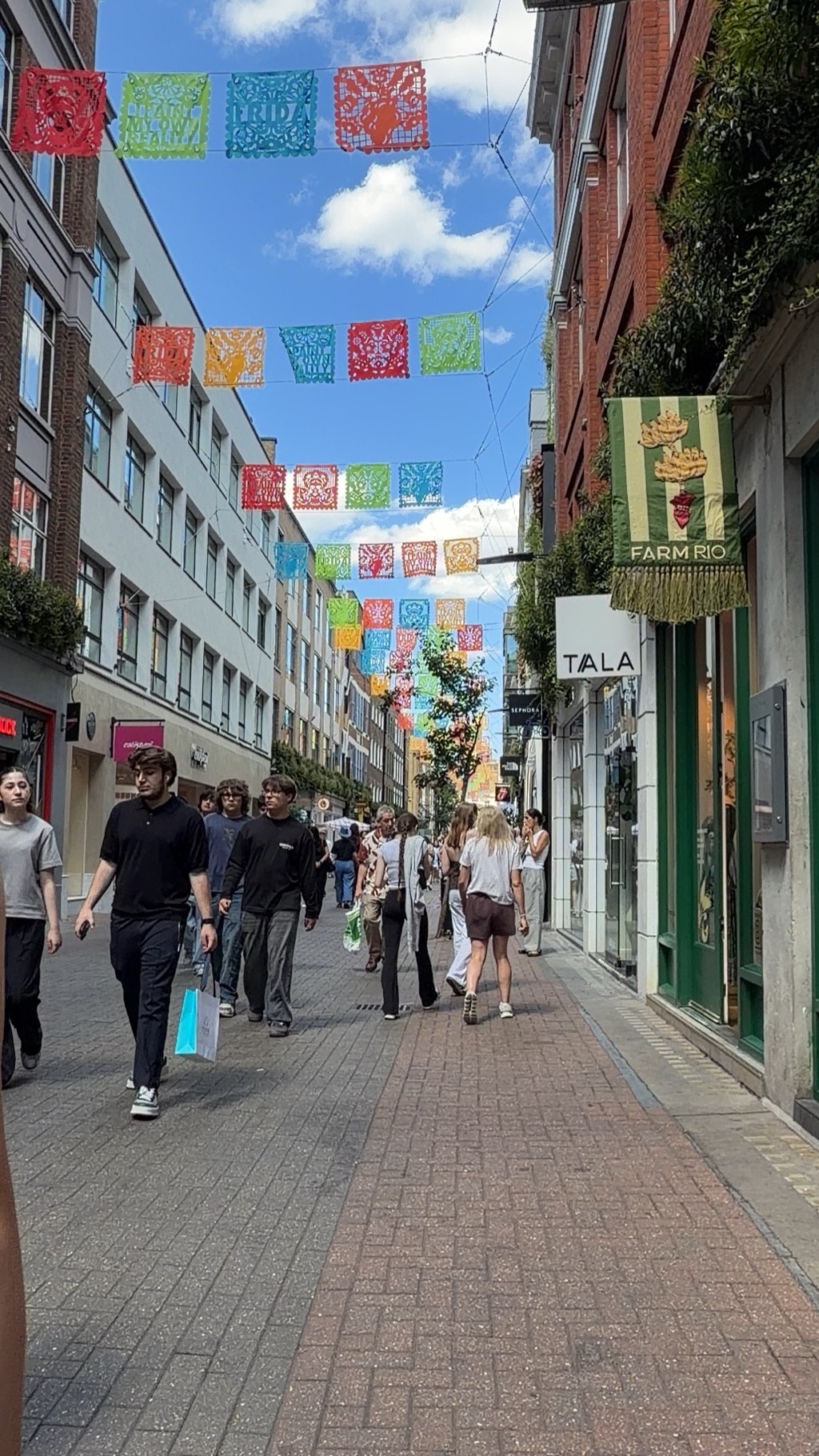 Looking down a narrow Carnaby Street side lane strung with rows of brightly colored papel picado bunting in red, green, orange, and blue against a blue sky with white clouds. Shoppers carrying bags walk along the brick-paved street past storefronts including a green-fronted Farm Rio shop with a fringed banner and a Tala sign