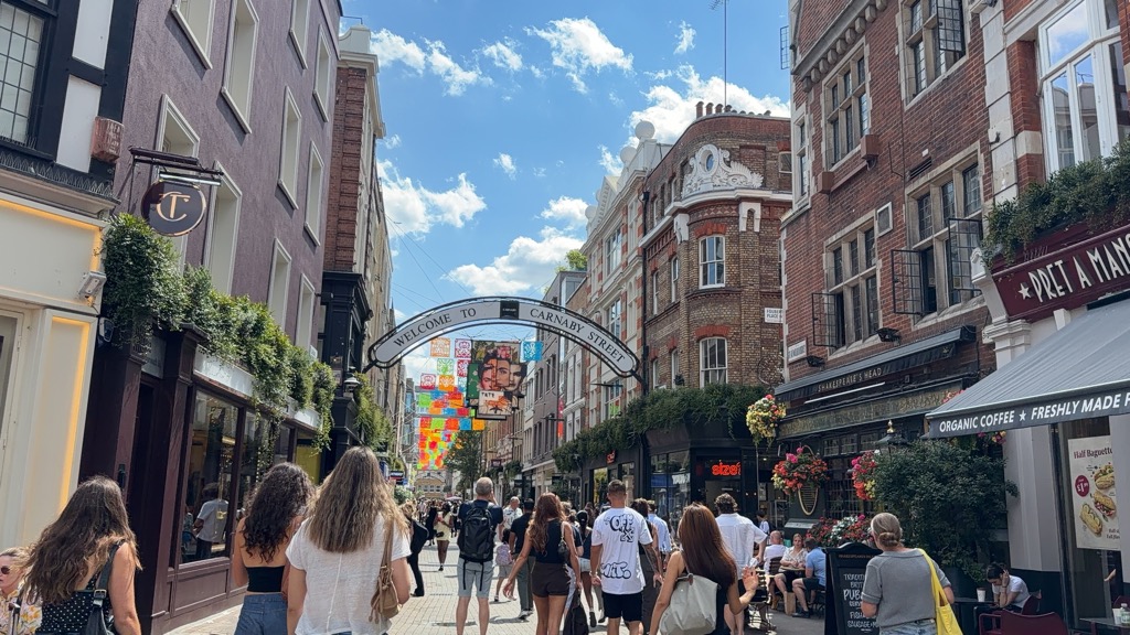 The pedestrianized Carnaby Street in London on a bright sunny day, with a white metal arch overhead reading 'Welcome to Carnaby Street' and a tall colorful banner installation of stacked painted panels and faces further down the street. Shoppers walk between brick and painted buildings lined with hanging plants, a Pret A Manger awning on the right, and a pub sign for Shakespeare's Head, under a blue sky with scattered clouds