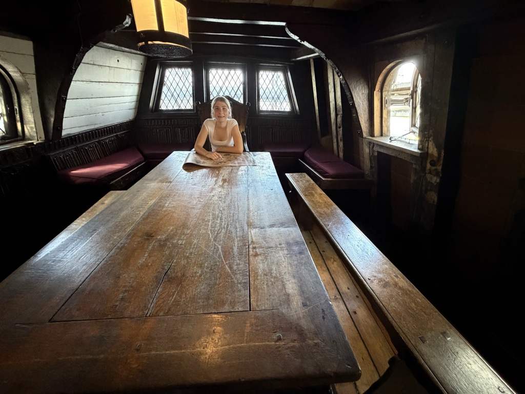 Below decks aboard the Golden Hinde replica galleon, showing a long scrubbed oak table running the length of a low timber-beamed cabin with benches and purple cushioned seating along the walls. A girl in a white top sits at the far end of the table beneath leaded diamond-pane windows, with a lantern hanging from the ceiling and small arched windows letting in daylight