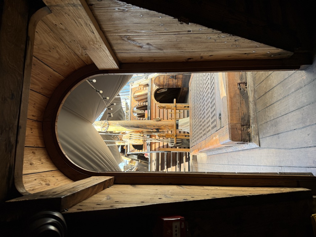 View through an arched wooden doorway aboard the Golden Hinde, looking out from a dark timber cabin onto the sunlit main deck. Beyond the doorway are a wooden grating hatch cover, coiled rigging around the mast, a ship's capstan, and stairs leading up, all sheltered under a white canvas awning strung with festoon lights