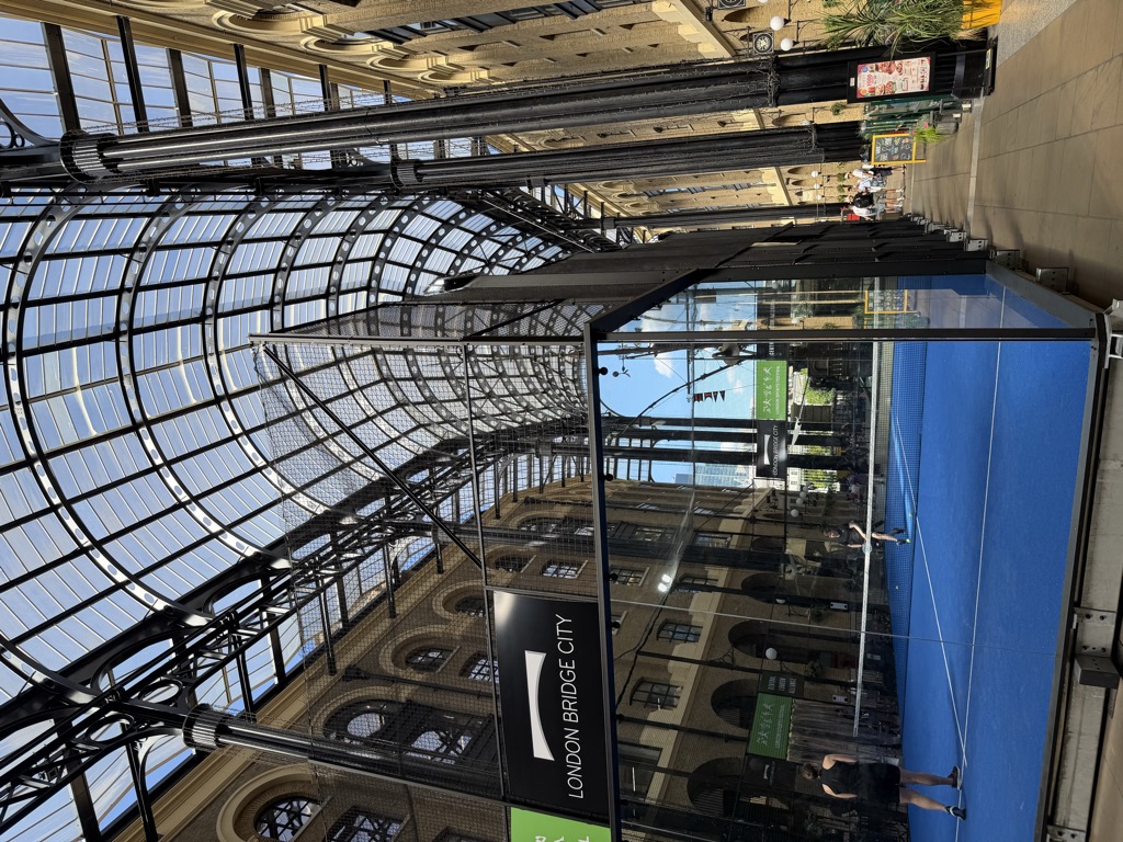 Inside Hay's Galleria at London Bridge City, a glass-roofed Victorian arcade with an arched lattice of black ironwork overhead and tall yellow brick buildings on both sides. A blue padel court enclosed in glass and netting has been installed on the ground floor, with two players mid-rally, and a black sign reading 'London Bridge City' hangs above