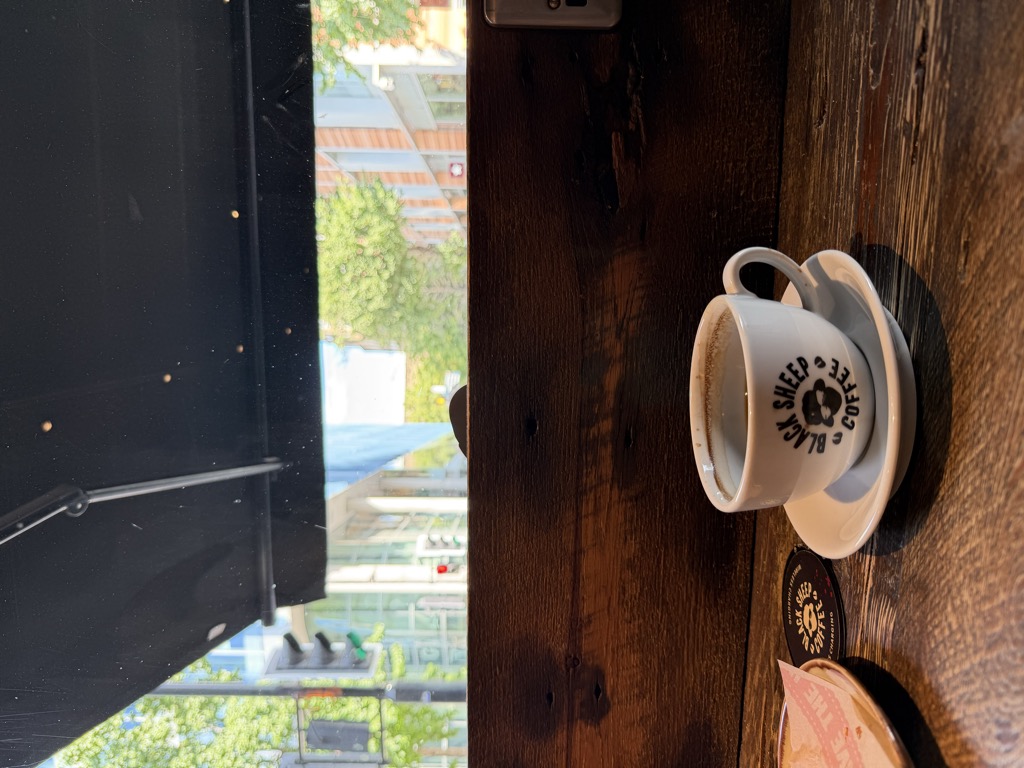 A white cup of black Americano coffee on a matching saucer, both stamped with the Black Sheep Coffee logo of a sheep's head in a circle, sitting on a dark reclaimed-wood counter beside a branded coaster and a pastry plate. Through the open window above the counter, a sunlit street with green trees, a traffic light, and glass office buildings is visible