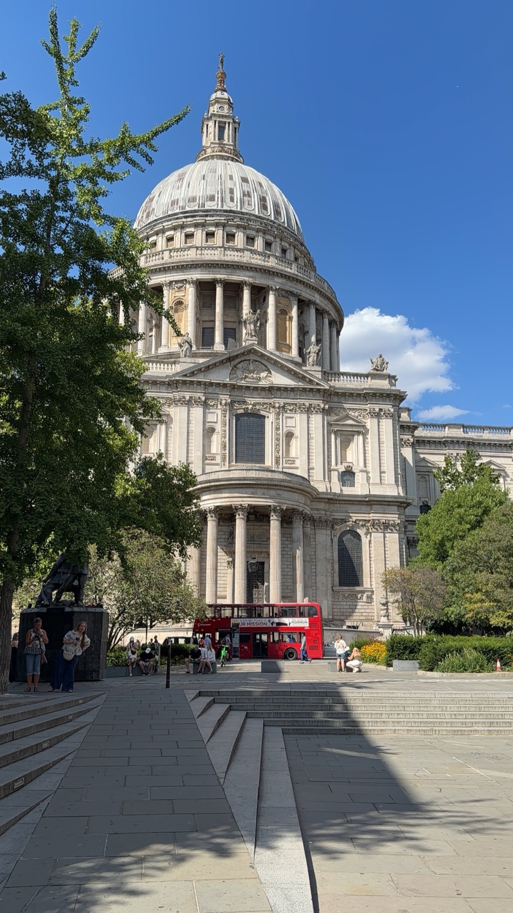 St Paul's Cathedral in London photographed from the gardens to the south, showing the full stone dome with its lantern and gold cross, the columned drum beneath, and the pedimented south transept entrance with statues along the roofline. A red double-decker bus is parked at the foot of the steps, visitors sit on benches in the shade, and a large tree frames the left side against a deep blue sky