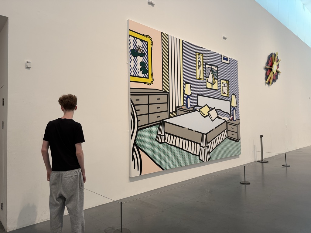 A large Roy Lichtenstein painting of a bedroom interior hanging on a white gallery wall at Tate Modern, rendered in his comic-book style with heavy black outlines and Ben-Day dots, showing a made bed with a striped valance, a chest of drawers, framed pictures, and table lamps in pastel pinks, greens, and blues. A teenage boy in a black t-shirt and grey sweatpants stands with his back to the camera looking at it, behind a low stanchion barrier