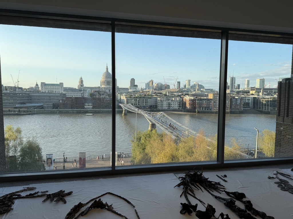 View from a gallery window at Tate Modern looking north across the River Thames in late afternoon light, with the Millennium Bridge crossing the water and pedestrians strung out along its span. The dome of St Paul's Cathedral rises above the City of London skyline, with construction cranes and office towers on the horizon. In the foreground, dark twisted sculptural forms are laid out across a white plinth