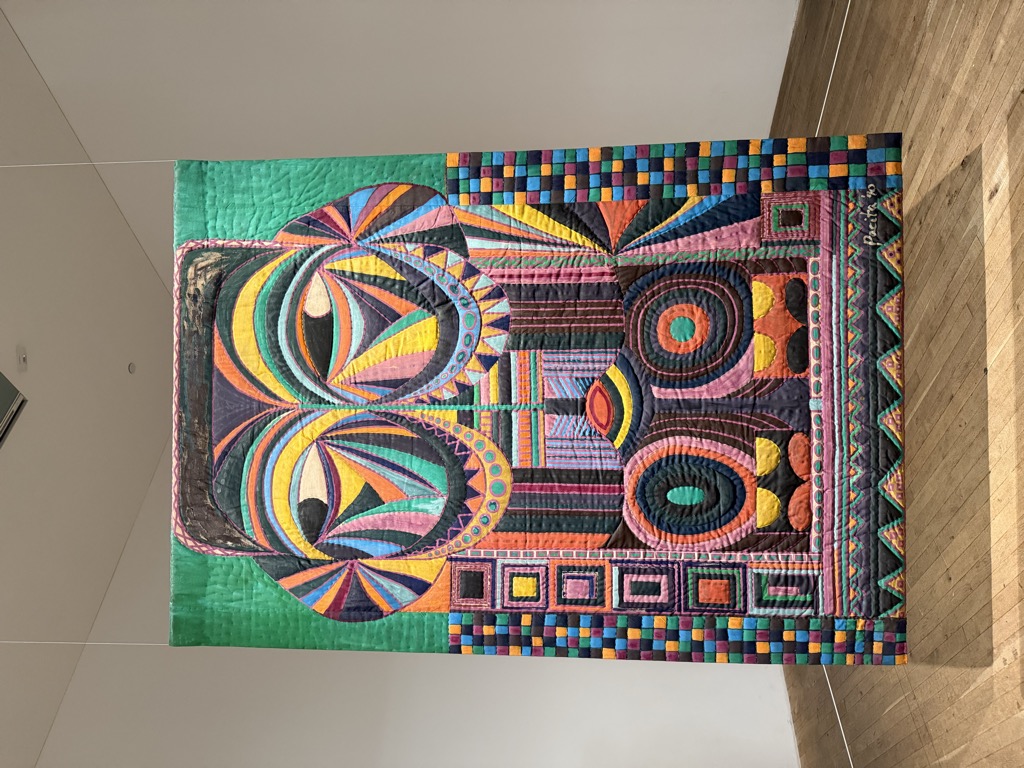 A large quilted textile artwork suspended by wires in a white gallery room at Tate Modern, hanging just above a pale wooden floor. The padded fabric panel depicts a stylized face in vivid stitched color, with two huge circular eyes made of radiating triangular segments in orange, yellow, blue, and purple, framed by a border of small multicolored squares and a band of zigzag triangles along the bottom