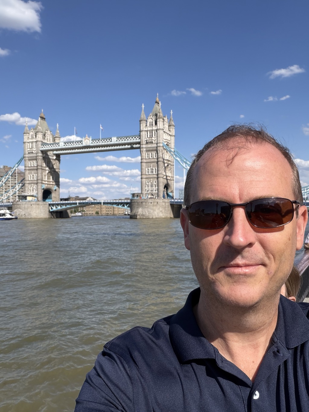 A selfie taken on the south bank of the River Thames with Tower Bridge filling the background on a clear sunny afternoon. The bridge's two Gothic stone towers with pointed turrets rise on either side of the pale blue high-level walkway, and the road deck and suspension chains span the green-brown water below, where a small white boat passes on the left. A man with short hair, wearing dark wraparound sunglasses and a navy polo shirt, stands at the right of the frame beside a metal railing, with a blue sky and scattered white clouds overhead