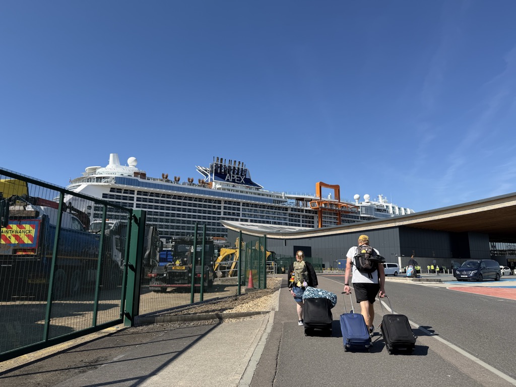 Walking toward the Horizon cruise terminal, the Celebrity Apex looms over the low modern terminal building on the right, its dark blue hull lettering reading APEX below the funnel, with the orange Magic Carpet platform parked partway up the side. A green mesh security fence runs along the left with construction plant, dump trucks, and a yellow excavator behind it. Two travelers with roller bags -- a man in a white t-shirt, dark backpack, and yellow cap, and a woman with a floral bag -- walk ahead of the camera down the roadway. High-vis-vested port staff and a black van are visible near the terminal doors. Cloudless blue sky.