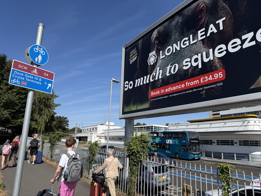 Looking up at the same blue Dock Gate 10 & Cruise Terminals sign, mounted below a blue shared cycle and pedestrian roundel on a metal pole, with a red SCN S4 route plate. To the right, a large billboard advertises Longleat with the tagline So much to squeeze and Book in advance from 34.95 pounds, showing a close-up of an elephant's eye. Below the billboard a teal Unilink double-decker bus passes on the road, and pedestrians with roller bags -- including a young man in a white t-shirt, grey backpack, and pink shorts -- walk along the sidewalk beside a metal railing.
