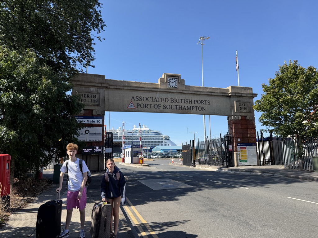 Two teenagers with roller bags stand in the road in front of the stone arch of Dock Gate 10 at the Port of Southampton. The arch is carved with ASSOCIATED BRITISH PORTS and PORT OF SOUTHAMPTON, with BERTH 102-109 on the left pier and GATE No 10 on the right, a clock face set into the top, and a Union Jack on a flagpole above. Through the archway a white cruise ship is visible at the berth beyond a gatehouse and security fencing. A red-headed boy in a white t-shirt and pink shorts and a girl in a navy hoodie and shorts pull black and tan suitcases. Bright sun, clear blue sky.