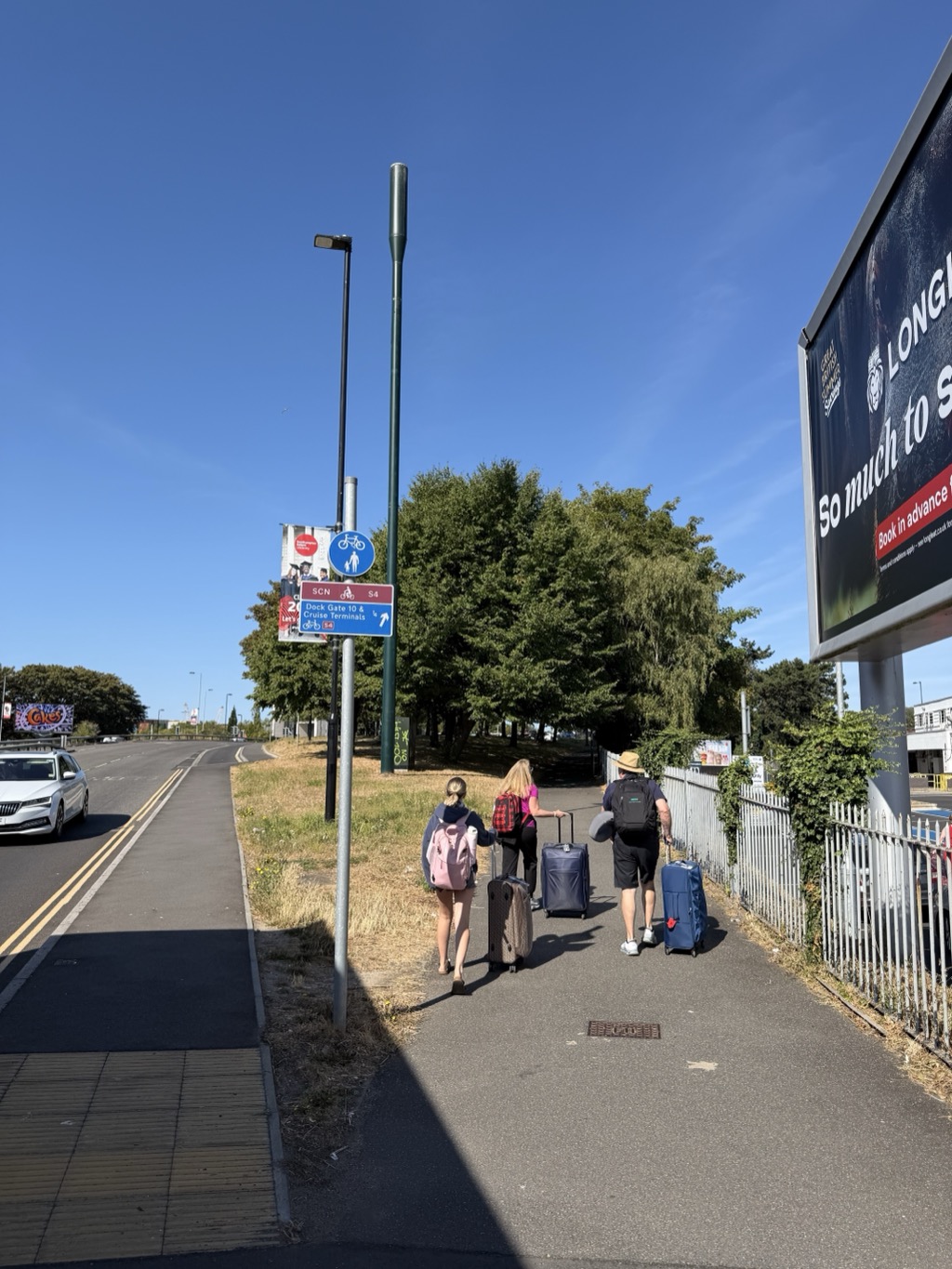 View up a sidewalk beside a road under a cloudless deep blue sky. In the middle distance, three people walking away from the camera with roller bags -- a young woman with a pink backpack and braided hair, a woman in a pink top with a plaid shirt tied around her waist, and a man in a straw hat, dark backpack, and shorts. A signpost in the foreground carries a blue cycle-and-pedestrian shared path roundel above a blue sign reading Dock Gate 10 & Cruise Terminals with an up-right arrow and a quarter-mile distance. A large billboard on the right shows a dark animal photo. Cars pass on the road at left, and the grass verge is scorched brown from summer heat.