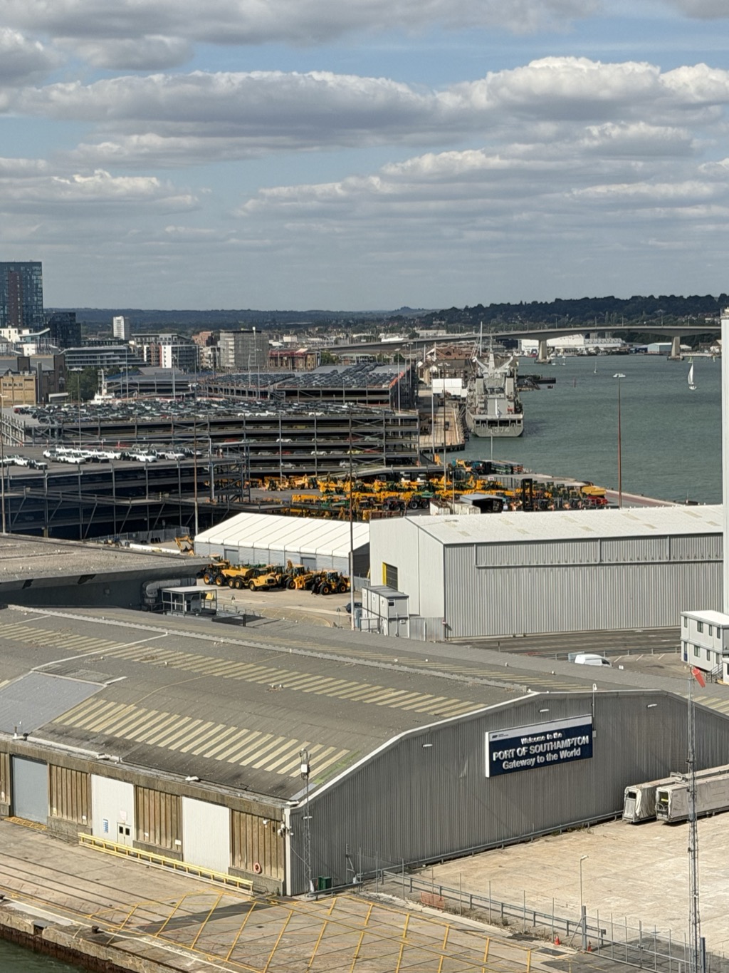 Elevated view from the ship over the Port of Southampton. In the foreground, a long grey warehouse with a navy sign reading Welcome to the PORT OF SOUTHAMPTON, Gateway to the World, beside an empty concrete quay marked with yellow lines. Behind it, a multi-story car park stacked with hundreds of new cars, rows of yellow construction machinery lined up on the dock, a white marquee tent, and a grey naval supply ship moored at a wharf. Beyond the water lies the far shore with a road bridge, wooded hills, and waterfront apartment blocks. Partly cloudy sky.
