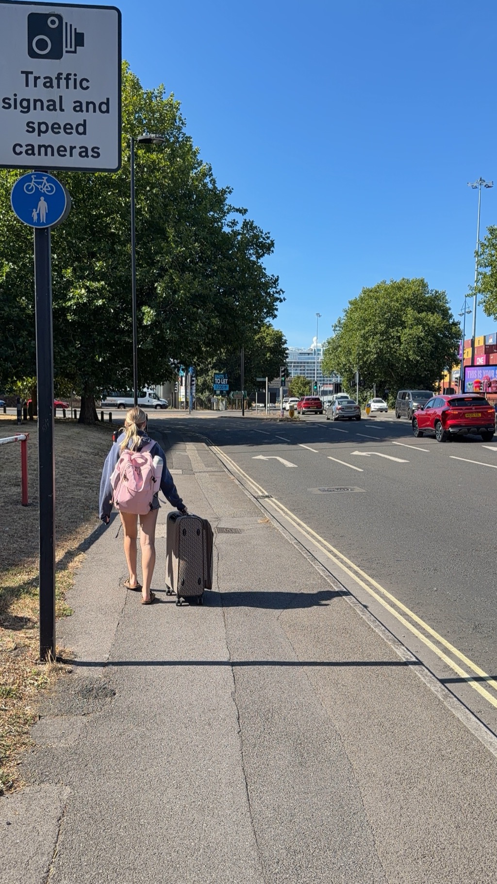 A young woman with a blonde ponytail, pink backpack, and a blue sweater draped over her shoulders walks away from the camera along a sidewalk pulling a tan patterned roller bag, wearing shorts and flip-flops. A tall sign in the left foreground reads Traffic signal and speed cameras above a blue shared cycle and pedestrian roundel. Plane trees line the left side; a wide multi-lane road with double yellow lines and a red SUV runs along the right. In the gap between the trees at the center of the frame, the white superstructure of a cruise ship is just visible above rooftops in the distance.