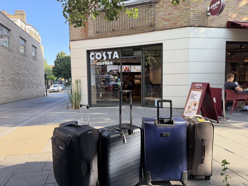 Four roller suitcases -- black, gunmetal, navy, and a patterned tan one -- lined up on a wide paved sidewalk outside a corner Costa Coffee in bright early morning sun. The shop occupies a brick and white-rendered building with a large plate glass window bearing the white COSTA COFFEE logo; a burgundy A-frame sign on the pavement advertises hazelnut creme delights. A man in a dark t-shirt sits at an outdoor table on the right. A yellow tactile paving strip runs along the left edge of the sidewalk and parked cars sit on the quiet street beyond.