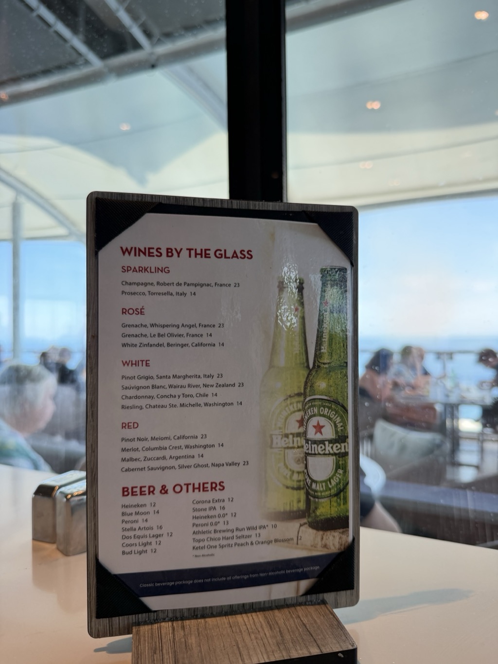 A laminated table-tent drinks menu standing on a white tabletop in a glass-walled shipboard venue. The card is headed WINES BY THE GLASS in red, with sections for Sparkling (Champagne, Robert de Pampignac, France 23; Prosecco, Torresella, Italy 14), Rose, White (Pinot Grigio, Santa Margherita, Italy 23; Sauvignon Blanc, Wairau River, New Zealand 23; Chardonnay, Concha y Toro, Chile 14; Riesling, Chateau Ste. Michelle, Washington 14), and Red (Pinot Noir, Meiomi, California 23; Merlot, Columbia Crest, Washington 14; Malbec, Zuccardi, Argentina 14; Cabernet Sauvignon, Silver Ghost, Napa Valley 23). A BEER & OTHERS block lists Heineken 12, Blue Moon 14, Peroni 14, Stella Artois 16, Dos Equis Lager 12, Coors Light 12, Bud Light 12, Corona Extra 12, Stone IPA 16, and Topo Chico Hard Seltzer 13, with fine print noting the classic beverage package does not include all offerings. Two green Heineken bottles are printed alongside. Behind the menu, salt and pepper shakers sit on the table and other passengers are seated at tables beside the windows, with flat blue ocean and sky beyond.