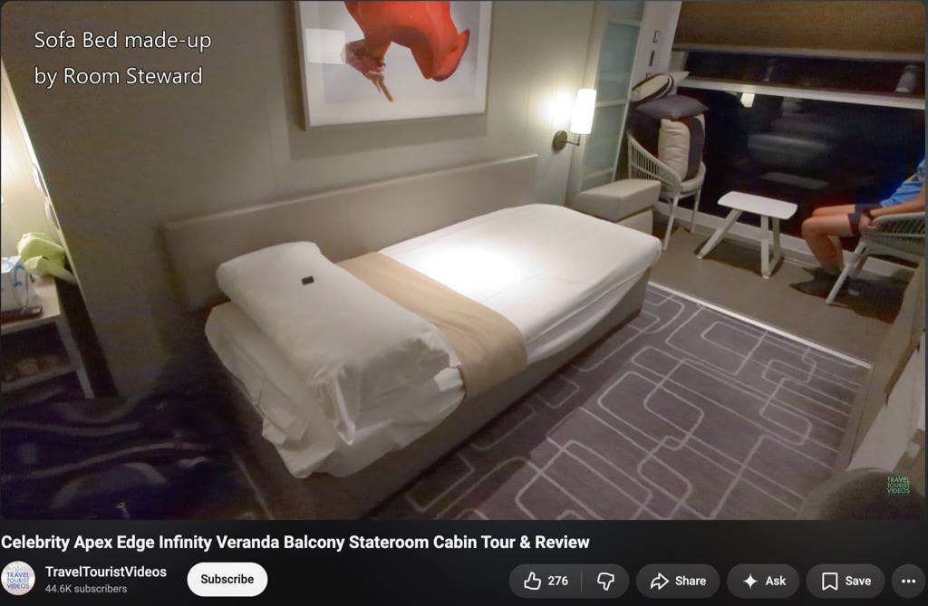 A screenshot of the YouTube video 'Celebrity Apex Edge Infinity Veranda Balcony Stateroom Cabin Tour & Review' by the channel TravelTouristVideos, which has 44.6K subscribers. The video frame shows an Edge Stateroom with the sofa converted into a made-up single bed, captioned in white text at the upper left 'Sofa Bed made-up by Room Steward'. The bed carries white sheets, a folded white duvet, and a tan runner across the foot, set against a padded headboard below a framed print of a red abstract form. A wall sconce with a rectangular shade glows beside it, and a grey rug patterned with white rounded rectangles covers the floor. At the right, the infinite veranda's glass wall is open to a darkened evening view, with a woven chair, an ottoman, and a small white side table on the veranda where a seated person in a blue top is partly visible. The YouTube interface below shows Subscribe, 276 likes, and Share, Ask, and Save buttons.