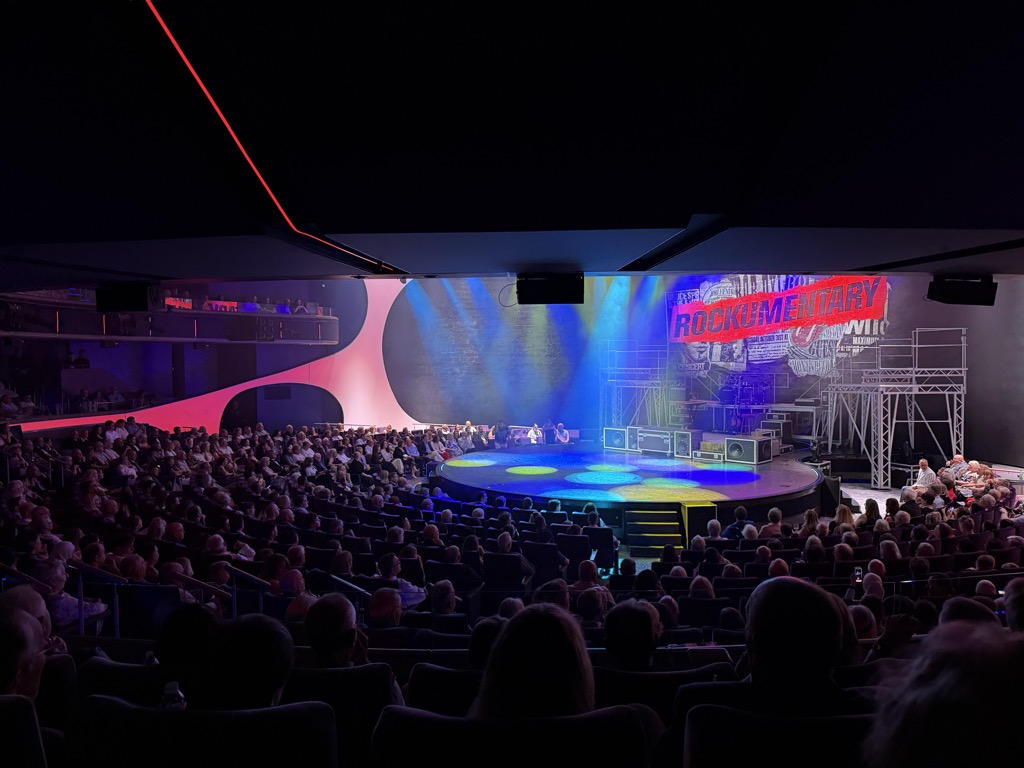 Looking down over a packed shipboard theater from the back rows. The round thrust stage in the center is lit in blue and yellow pools of light with a haze of theatrical smoke, ringed by stacked speaker cabinets and scaffolding towers. A large projection at the upper right reads ROCKUMENTARY in red block letters over a collage backdrop. Curved pink and white sculptural panels frame the stage at left, a red LED strip traces the dark ceiling, and an upper balcony level is visible along the left wall. Several hundred seated audience members fill the dark auditorium in silhouette.