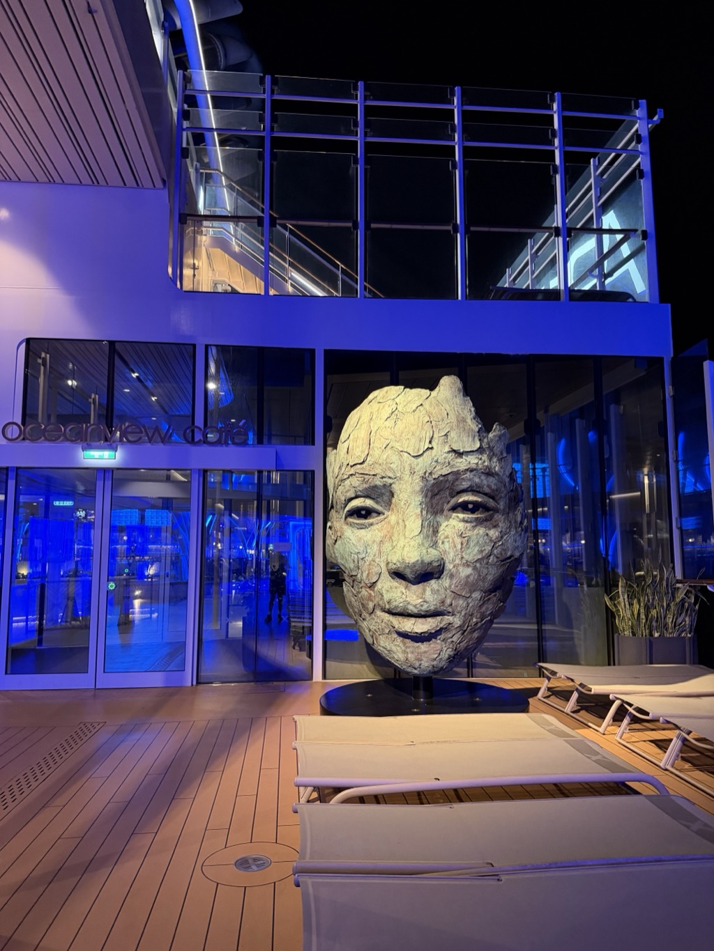 Night shot on the pool deck outside the Oceanview Cafe, whose backlit lowercase sign glows above the glass entrance doors at left. The ship's superstructure is washed in deep blue and violet uplighting, with a glass-railed upper deck above. In the center, a large sculpture of a human head rests on a low black plinth -- a pale, weathered, cracked-clay face with heavy-lidded eyes and a closed mouth, lit warmly against the blue. Empty cream lounge chairs sit on the warm-toned teak deck in the foreground, a potted plant stands to the right, and the sky beyond is black.