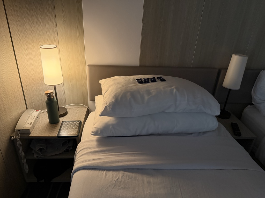 A cruise ship stateroom bed after turndown service, shot in warm low light. Two plump white pillows are stacked against a padded grey headboard with four small dark foil-wrapped chocolates lined up neatly on top, and the white duvet is smoothed flat across the bed. A cylindrical shaded lamp glows on the light wood nightstand at left beside a white cabin telephone, a dark green insulated water bottle, and a small tablet propped in a stand; a folded towel sits on the shelf below. A second matching lamp stands at right, with a black TV remote on the far nightstand. The cabin walls are finished in vertical pale wood veneer panels.