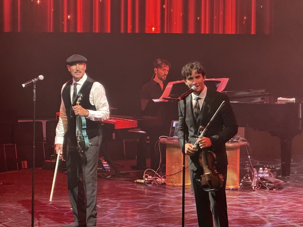 Two violinists performing on a dark shipboard theater stage lit in deep red, with a band of vertical red light strips across the backdrop above them. At left, an older man in a flat cap, black waistcoat, white shirt, tie, and grey trousers holds a violin and bow at his sides, a moustache and slight smile on his face. At right, a much younger performer in a dark three-piece suit and patterned tie sings into a microphone, curly dark hair, holding his violin down by his hip. Behind them a keyboardist in a black shirt plays a red keyboard beside a grand piano, with sheet music stands, cables, and water bottles scattered across the stage floor.