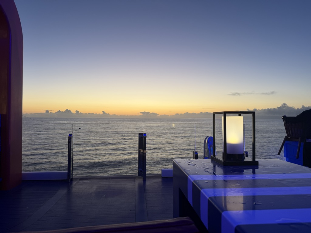 Dusk from an open-air bar at the stern of the ship, looking out over a calm Atlantic. The sky graduates from deep blue-violet overhead to a broad band of gold and amber at the horizon, with a low line of small dark clouds catching the last light and a faint glint of sun reflected on the water. In the foreground at right, a grey bar counter beaded with water is lit from beneath in electric blue, with a square black-framed lantern holding a glowing white pillar candle standing on it. Glass windbreak panels and a curved red-lit wall frame the left side, and dark wicker chairs sit at the right edge of the deck.