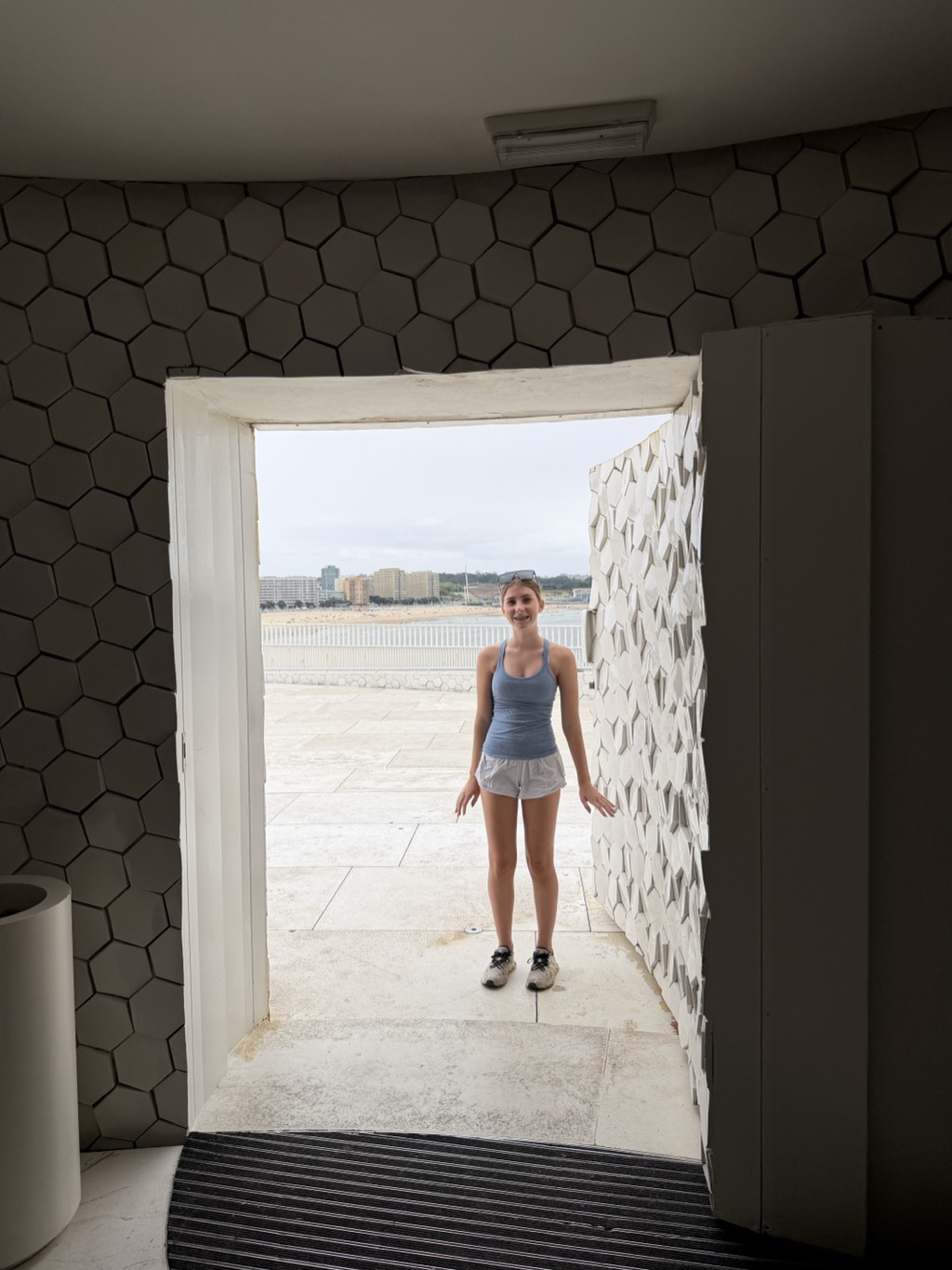Shot from inside the Leixoes terminal, looking out through an open doorway framed by walls tiled edge to edge in matte olive-grey hexagons. Siena stands centered in the bright doorway on the pale stone terrace outside, smiling at the camera, wearing a light blue ribbed tank top, grey athletic shorts, white sneakers, and sunglasses pushed up on her head, arms held slightly out from her sides. The open door leaf on the right is faced in white faceted panels. Beyond her, a white railing runs along the terrace edge, with the beach, the low white and tan apartment blocks of the waterfront, and a flat grey overcast sky in the distance.