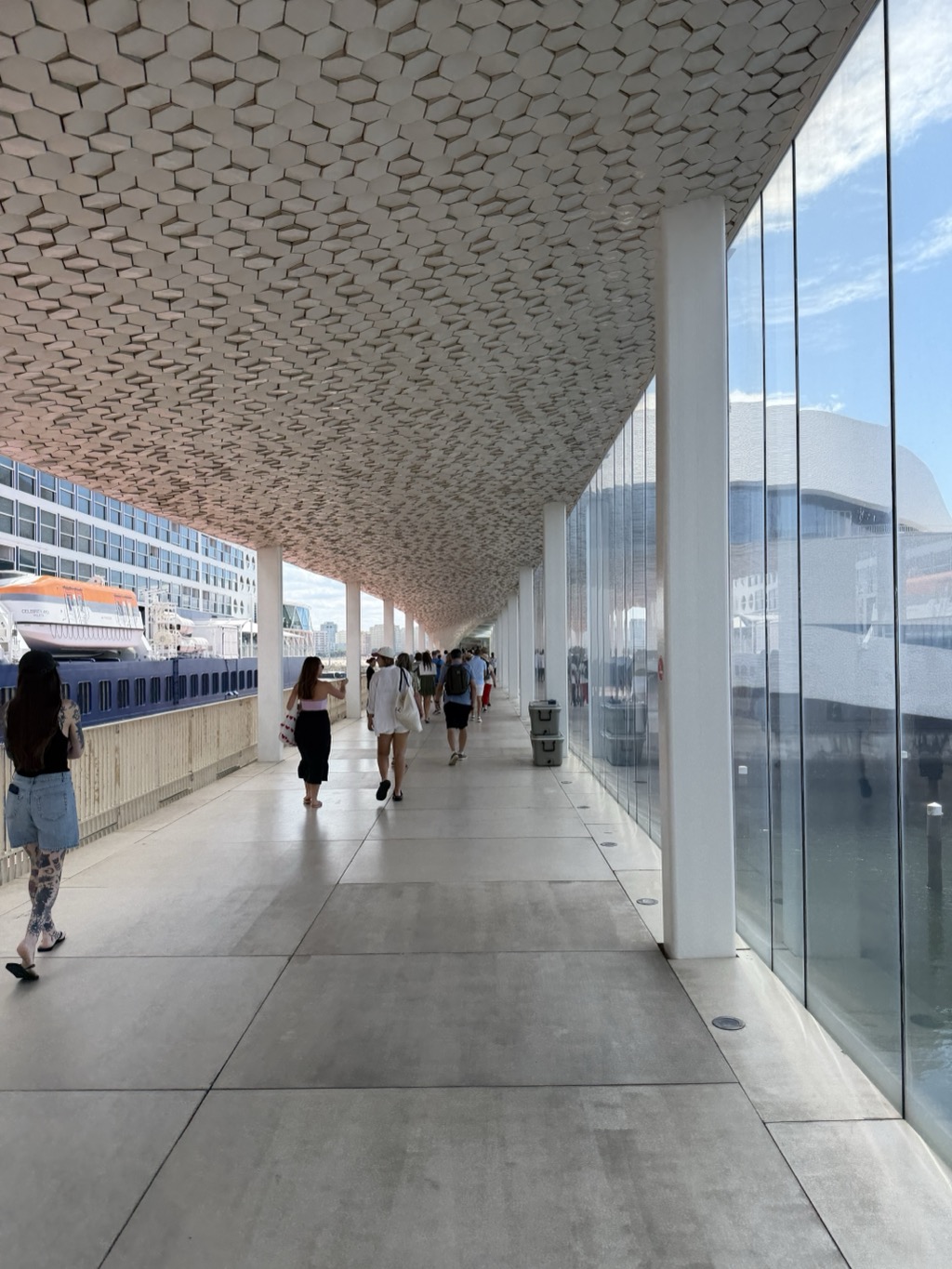 A long covered walkway at the Leixoes cruise terminal, its underside clad in thousands of small cream hexagonal tiles that ripple across the curved ceiling. Slender white columns line the right side beside a full-height glass wall reflecting the terminal's white curved facade and the water beyond. Passengers walk away from the camera down the pale concrete deck -- a woman in a black tank top and denim shorts with long dark hair and tattooed legs in the foreground, others further on in sun hats and backpacks. To the left, the white hull and orange Celebrity lifeboats of the ship are visible over a wooden railing. Bright blue sky with scattered cloud.