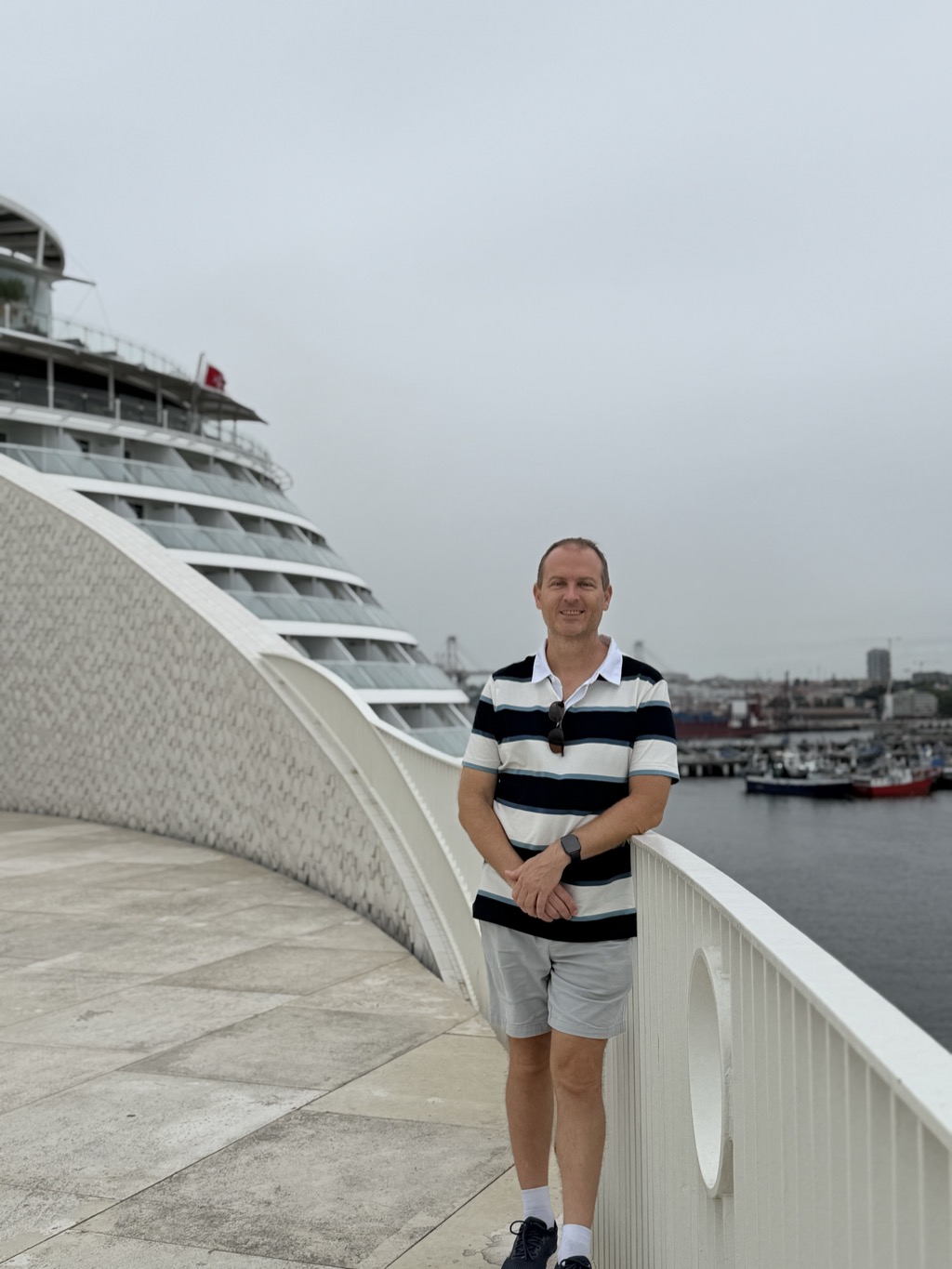 Brian stands on the curved white terrace of the Leixoes cruise terminal, leaning back against its low white railing, smiling at the camera. He wears a horizontally striped polo shirt in navy, white, and pale blue, light grey shorts, a dark watch, navy sneakers with white socks, and sunglasses hooked on his collar. Behind him at left, the tiled sweep of the terminal's curved wall leads the eye to the Celebrity Apex moored alongside, its tiered glass-fronted balcony cabins stacked above the waterline and a red flag flying from the upper deck. To the right, small fishing boats sit moored in the harbor with port cranes, container stacks, and the town skyline beyond under a flat grey sky.