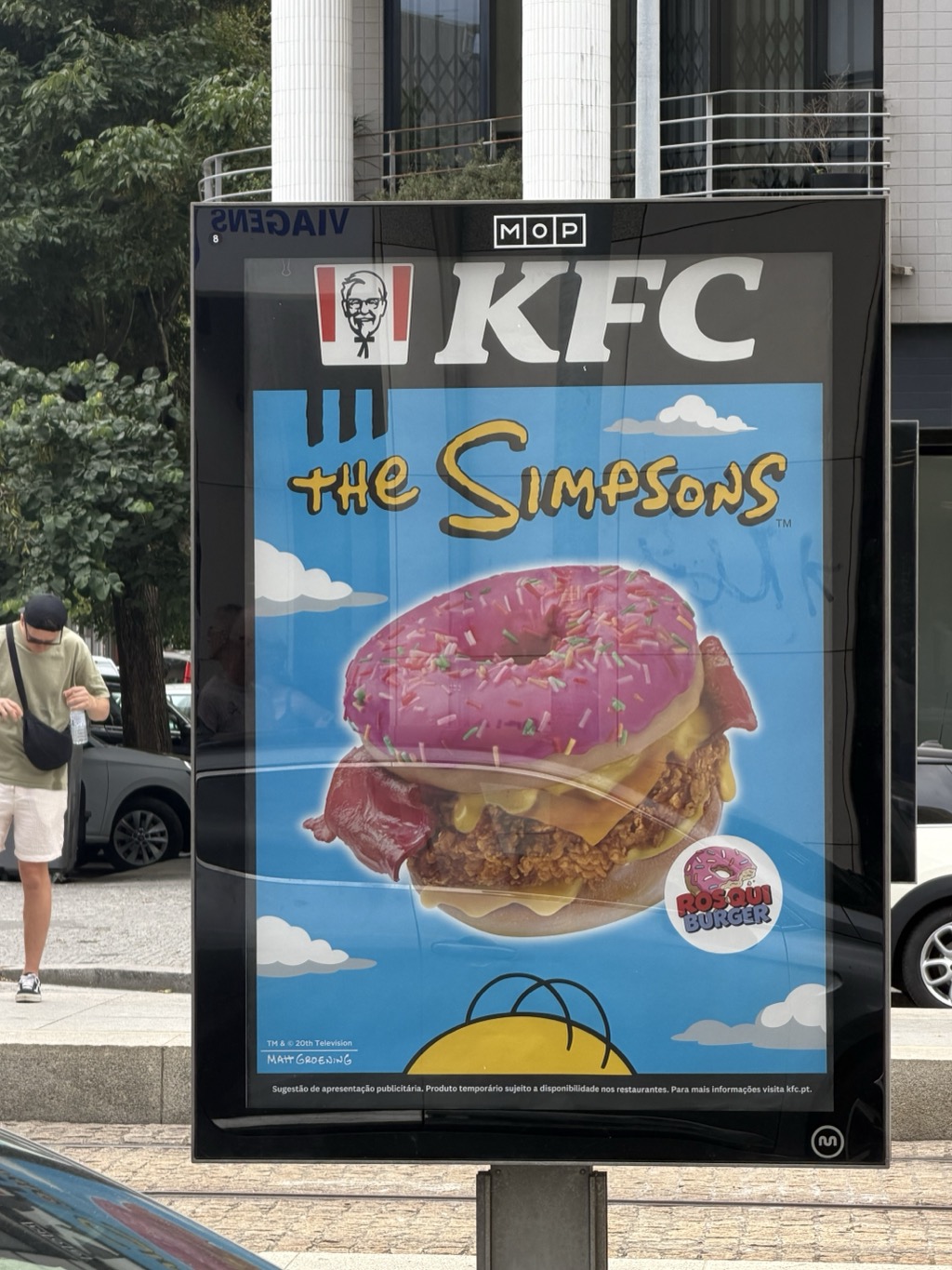 A glossy backlit street advertising panel on a Matosinhos sidewalk showing a KFC promotion tied to The Simpsons. The Colonel Sanders logo and KFC wordmark sit at the top; below, against a cartoon blue sky with white clouds, a photographed burger is topped with a pink sprinkled donut in place of the bun, stacked with fried chicken, bacon, and melted cheese. A yellow badge reads ROSQUI BURGER, and Homer Simpson's head appears in silhouette at the bottom. Portuguese fine print reads Sugestao de apresentacao publicitaria. A man in a green t-shirt, white shorts, dark cap, and crossbody bag walks past at the left, with parked cars and a modern concrete building behind.