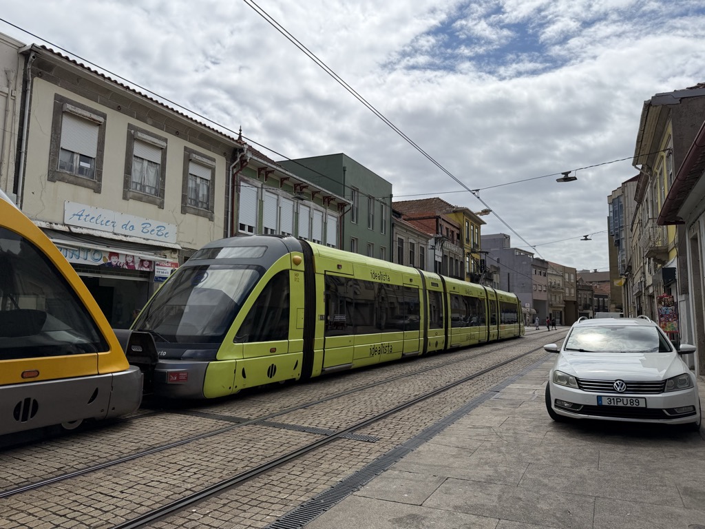 A lime-green and yellow Metro do Porto light rail tram stopped on cobbled tram tracks running down a narrow Matosinhos street, with the nose of a second yellow tram at the left edge of the frame. The carriages carry idealista branding along their flanks. Older two- and three-story buildings line both sides -- weathered stucco and tile facades with roller shutters, one bearing a hand-lettered sign reading Atelier do BeBe. Overhead catenary wires cross the frame against a heavily overcast grey and white sky. A white VW estate car with the Portuguese plate 31PU89 is parked on the granite-paved sidewalk at right.