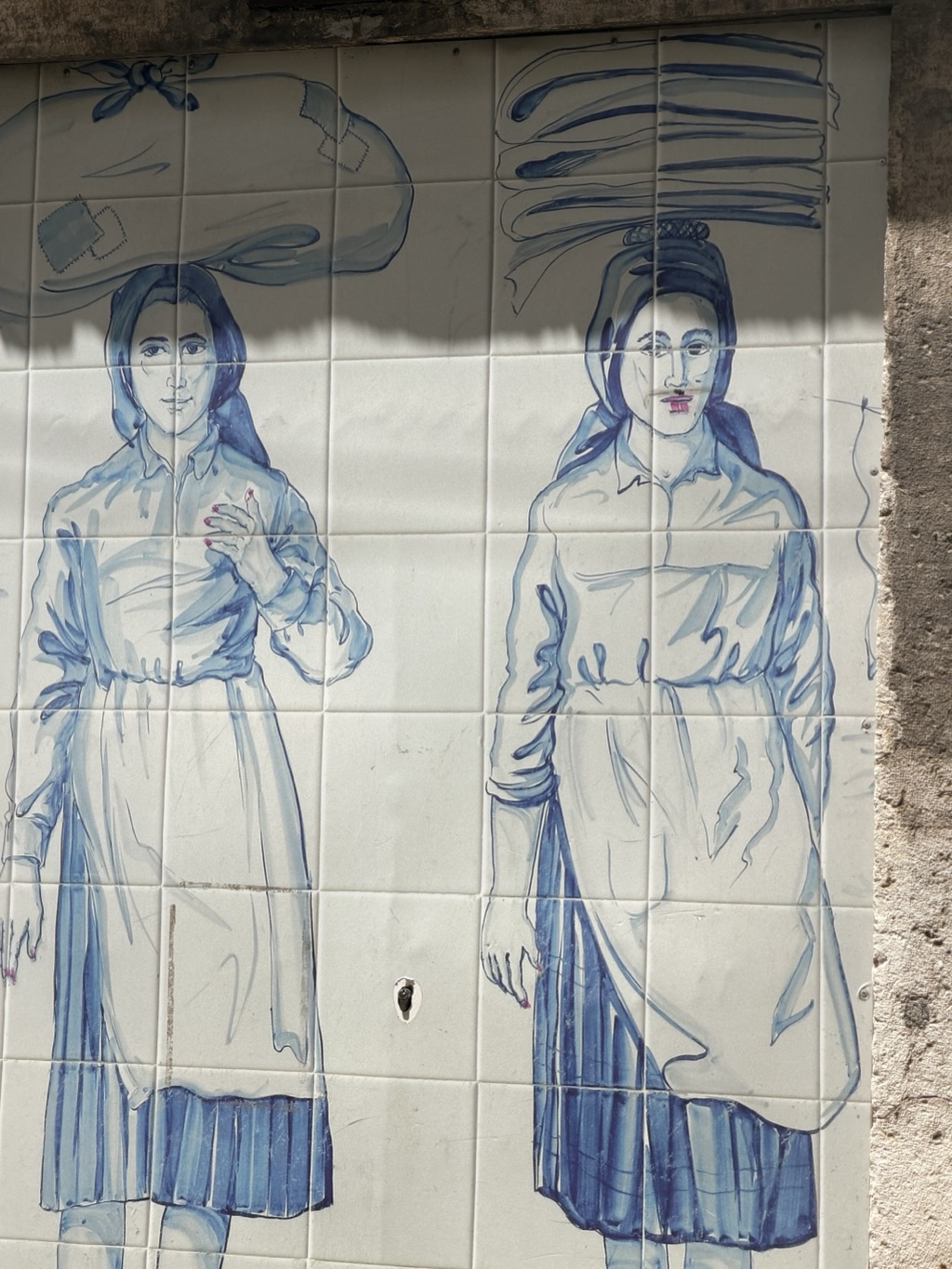A large blue-and-white azulejo tile panel on an exterior wall, painted in loose brushwork with two life-sized women in traditional dress. Each wears a headscarf, a long-sleeved blouse, an apron over a pleated skirt, and carries a load balanced on her head -- a broad cloth-wrapped bundle on the left figure, a stack of flat baskets or trays on the right. Both are painted mostly in cobalt blue washes on white tile; the right-hand figure has been vandalized with a pink mouth scribbled over her lips, and both have small pink marks added at the fingertips. The tiles are grimy and cracked in places, with a spray-painted grey band across the upper section and rough grey stone wall visible at the right edge.