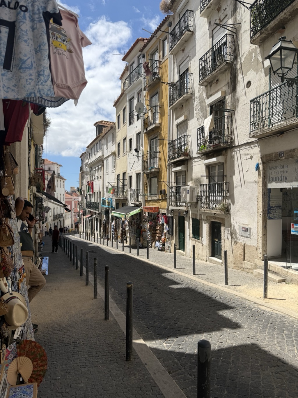 A narrow cobbled street in Alfama on a bright afternoon. Tall, weathered four- and five-story buildings line both sides in white, cream, and mustard yellow, with wrought-iron balconies, satellite dishes, laundry hung out to dry, and a Portuguese flag. Souvenir shops crowd the street level -- racks of t-shirts, bags, straw hats, and painted fans hang in the immediate left foreground, and a green-awninged stall further down displays magnets and postcards. A man in a dark vest and khakis leans against the shopfront at the left, talking on his phone. A shopfront at the right is signed lavandaria self service laundry at number 115. A line of black bollards separates the sidewalk from the granite-paved roadway, which is empty of cars.
