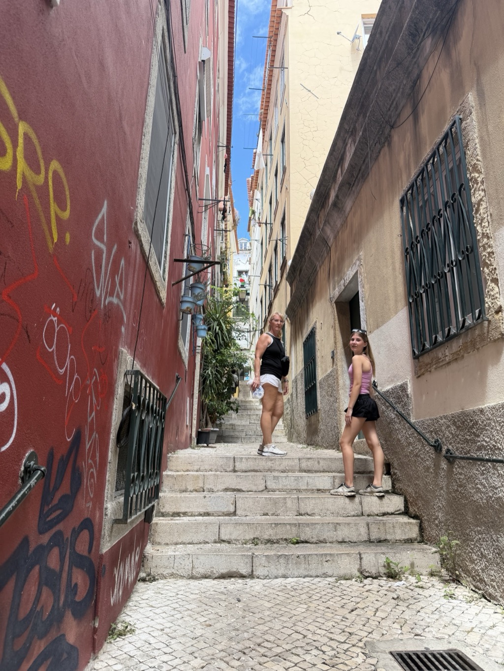 A narrow stepped alley climbing between tall buildings in Alfama. The left wall is deep red and heavily covered in layered graffiti tags in yellow, white, red, and black, with a barred window set into it. The right wall is bare cream stucco with a green-barred window and an open doorway. Sherri stands midway up the stone steps in a black tank top, white shorts, and white sneakers, holding a water bottle and turning back toward the camera; Siena stands a few steps below on the right in a pink tank top, black skirt, and sneakers, also looking back. Higher up, potted plants hang on brackets from the left wall and laundry is strung across the gap between the buildings, with a strip of bright blue sky visible far above.