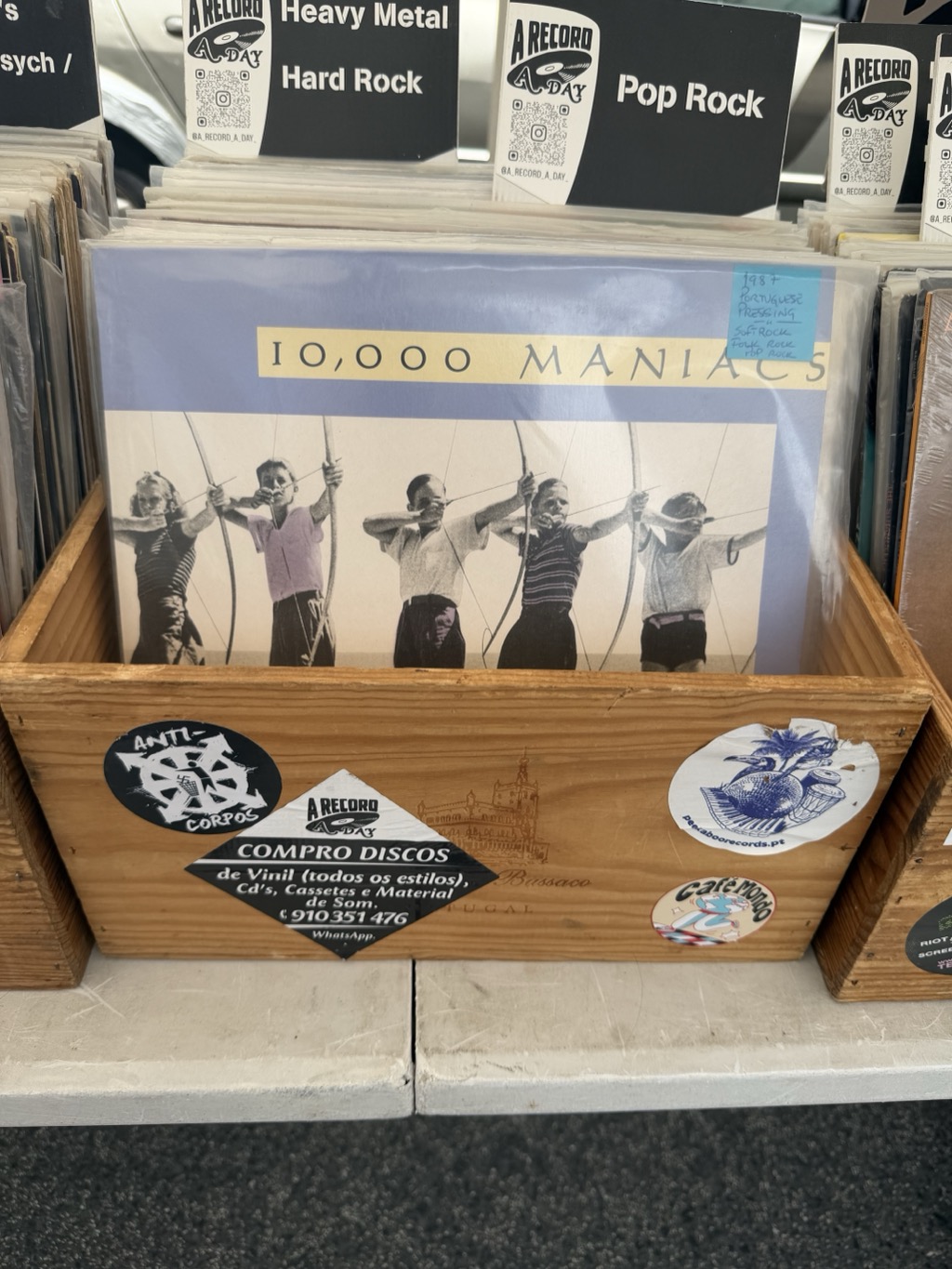 A wooden crate of second-hand vinyl records on a stall table at the Feira da Ladra flea market. The record facing forward is 10,000 Maniacs, its sleeve showing a black-and-white photograph of six children and adults drawing bows in an archery line against a cream background, with a lavender border. A handwritten blue sticker on the sleeve reads 1987 Portuguese Pressing, Soft Rock, Folk Rock, Pop Rock. The crate front is covered in stickers -- a black Anti-Corpos punk logo, a diamond-shaped A Record A Day card reading COMPRO DISCOS de Vinil (todos os estilos), CD's, Cassetes e Material de Som with the phone number 910 351 476 and WhatsApp, a blue Café Mondo sticker, and a discosboorecords.pt sticker. Black-and-white genre divider cards stand above the racks behind, labelled Psych, Heavy Metal, Hard Rock, and Pop Rock.