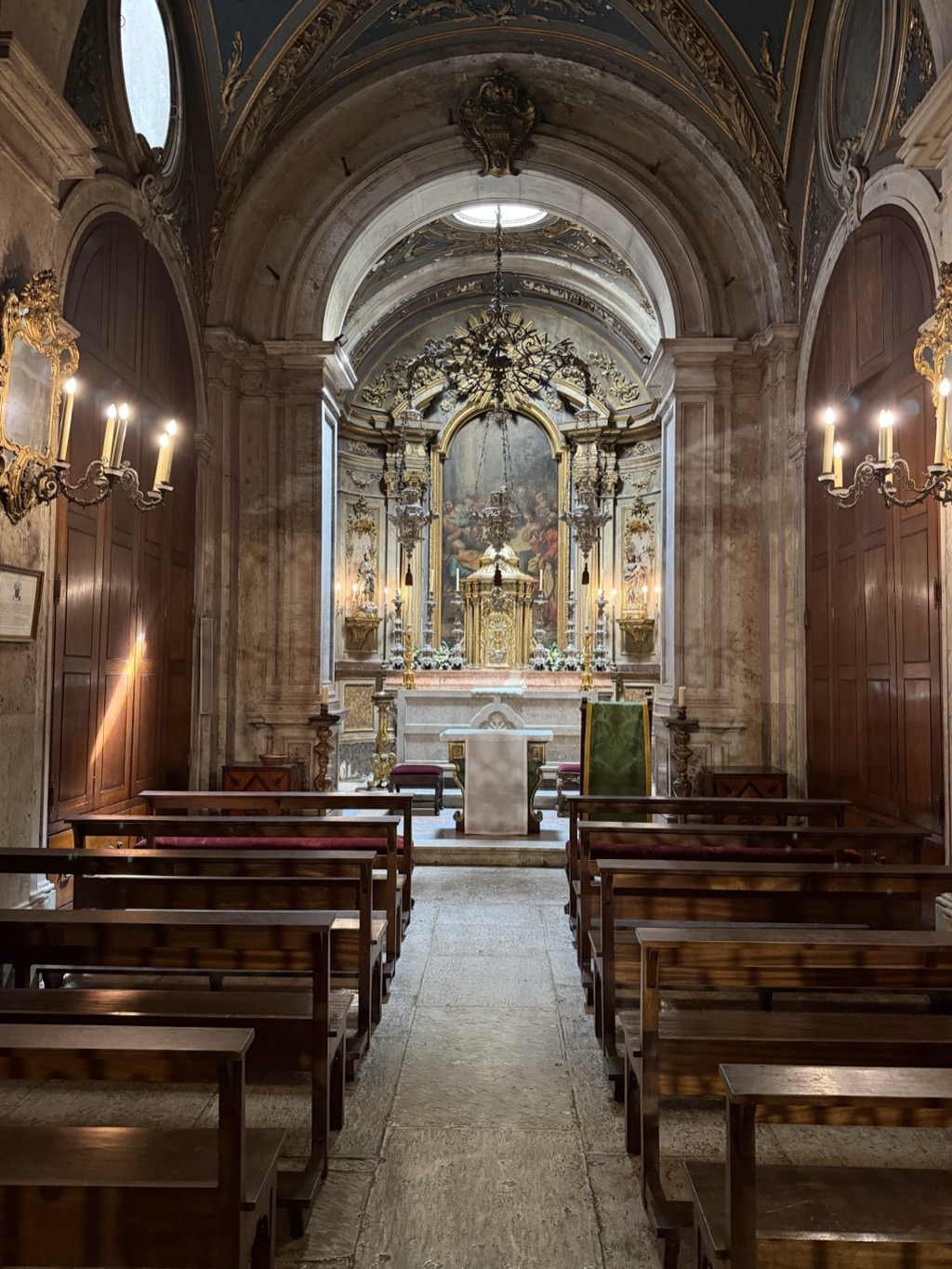 The interior of a small baroque church, viewed down the center aisle between rows of dark wooden pews. A barrel-vaulted ceiling with gilded plasterwork leads to an apse containing a heavily gilded altarpiece: twisting columns and carved scrollwork frame a large oil painting of a gathering of figures, with an ornate golden tabernacle on the marble altar below and silver candlesticks and flower arrangements on either side. A large brass chandelier hangs on a chain from the vault above the aisle. Wall sconces with electric candle bulbs flank tall wooden panelled doors at left and right, a green-draped celebrant's chair stands beside the altar, and worn stone flags pave the aisle.