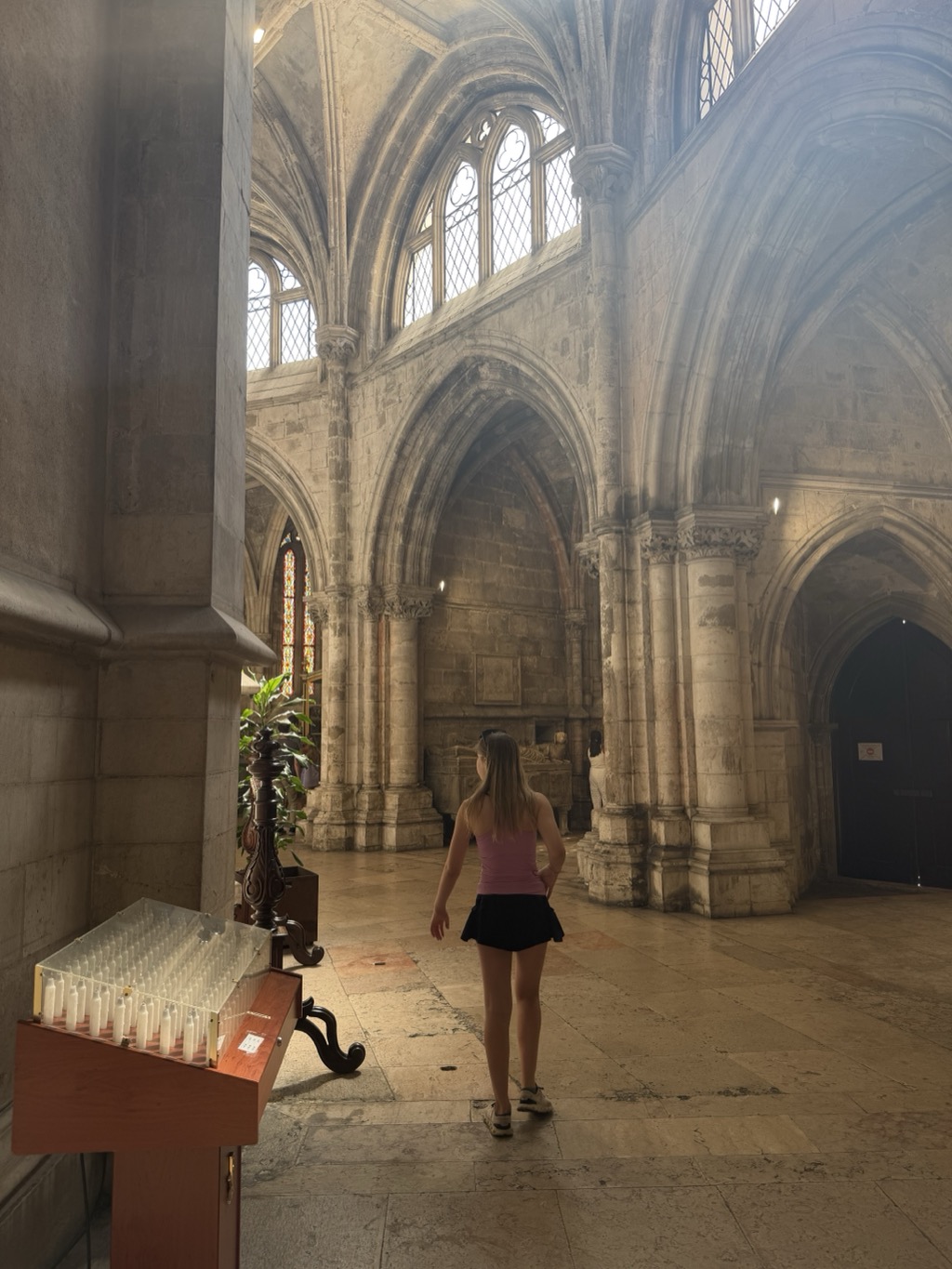 Inside Lisbon Cathedral, looking across the ambulatory at a soaring Gothic interior of pale limestone. Pointed arches spring from clustered columns with carved capitals, and tall lancet windows with leaded glass fill the clerestory above, sending shafts of hazy light across the space. Siena walks away from the camera across the worn stone floor, wearing a pink tank top, short black skirt, and sandals, glancing back to her right. In the left foreground a wooden votive candle stand holds rows of unlit white candles behind glass. A carved stone tomb chest and a potted palm stand along the far wall, with a jewel-toned stained glass window glowing in the aisle beyond.