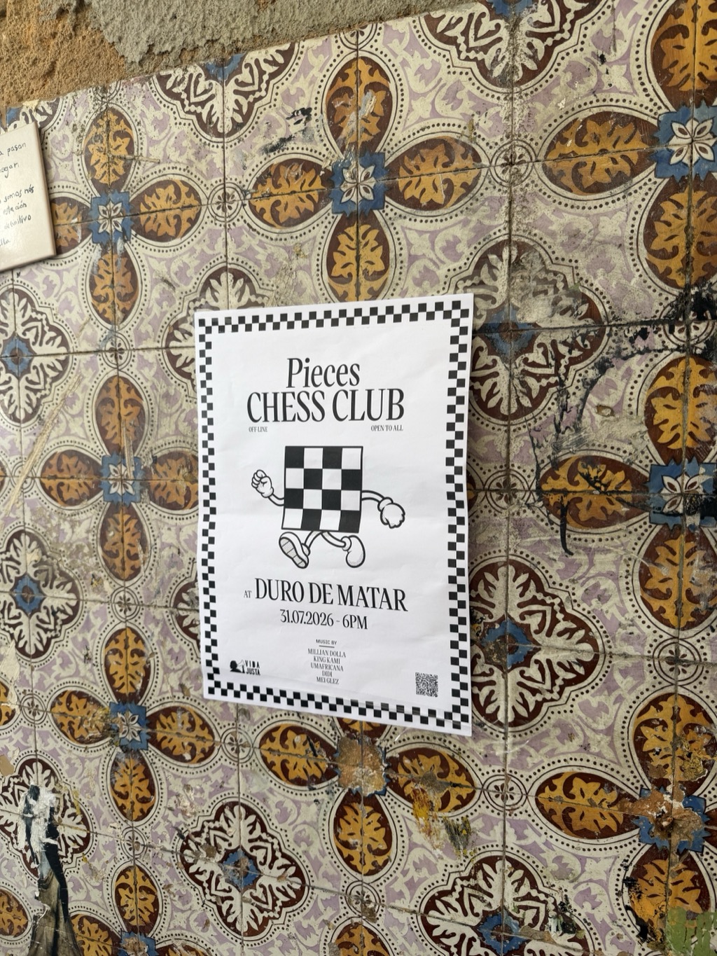 A photocopied white poster taped to a Lisbon wall entirely faced in old azulejo tiles patterned with repeating ochre, brown, blue, and cream floral medallions, chipped and scuffed with age. The poster has a black-and-white checkerboard border and reads Pieces CHESS CLUB, with OFF-LINE at the left and OPEN TO ALL at the right. Below is a cartoon of a checkerboard chess board with arms, legs, and a raised fist walking along. The text continues AT DURO DE MATAR, 31.07.2026 - 6PM, then MUSIC BY with a list of names -- Millian Dolla, King Kami, Umafricana, Didi, Mei Glez -- with a small Vida Justa logo at bottom left and a QR code at bottom right. A handwritten note in Spanish is taped to the wall at the upper left.