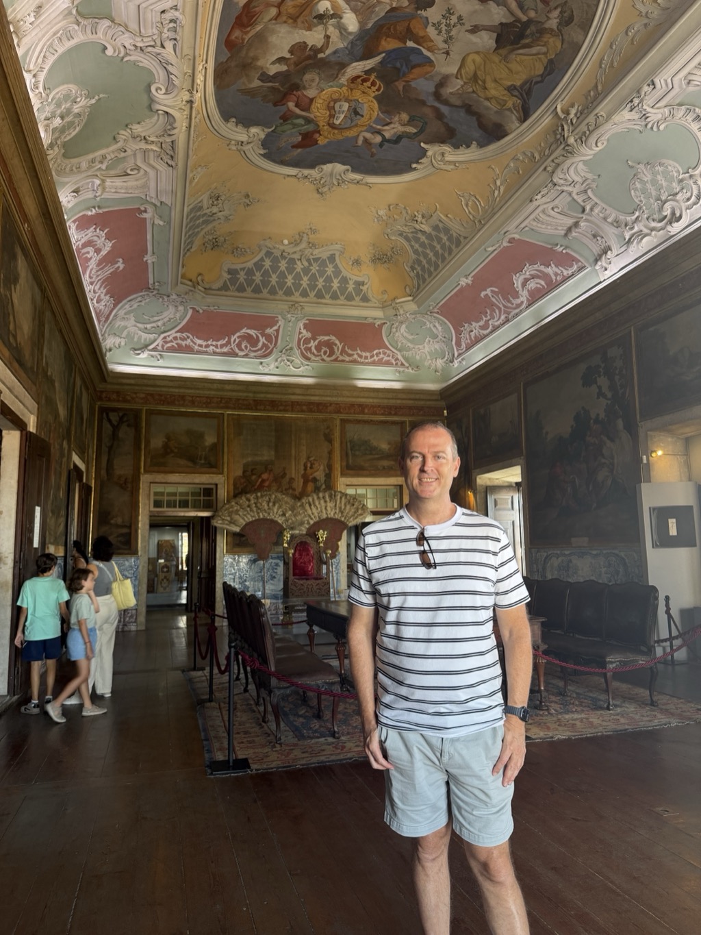Brian stands in the middle of a long, ornately decorated hall, smiling at the camera in a white t-shirt with thin navy horizontal stripes, pale blue shorts, a dark watch, and sunglasses hooked at his collar. Above him the coved ceiling is elaborately painted -- a central fresco of robed figures and cherubs floating on clouds around a gilded royal coat of arms, framed by white rococo plasterwork scrolls against panels of sage green, dusty pink, and ochre. The walls carry large gilt-framed landscape paintings and blue-and-white azulejo tilework, with an ornate carved and gilded canopy over a red-upholstered ceremonial chair in the middle distance. Dark polished floorboards run the length of the room, and a family of visitors moves through a doorway at the left.