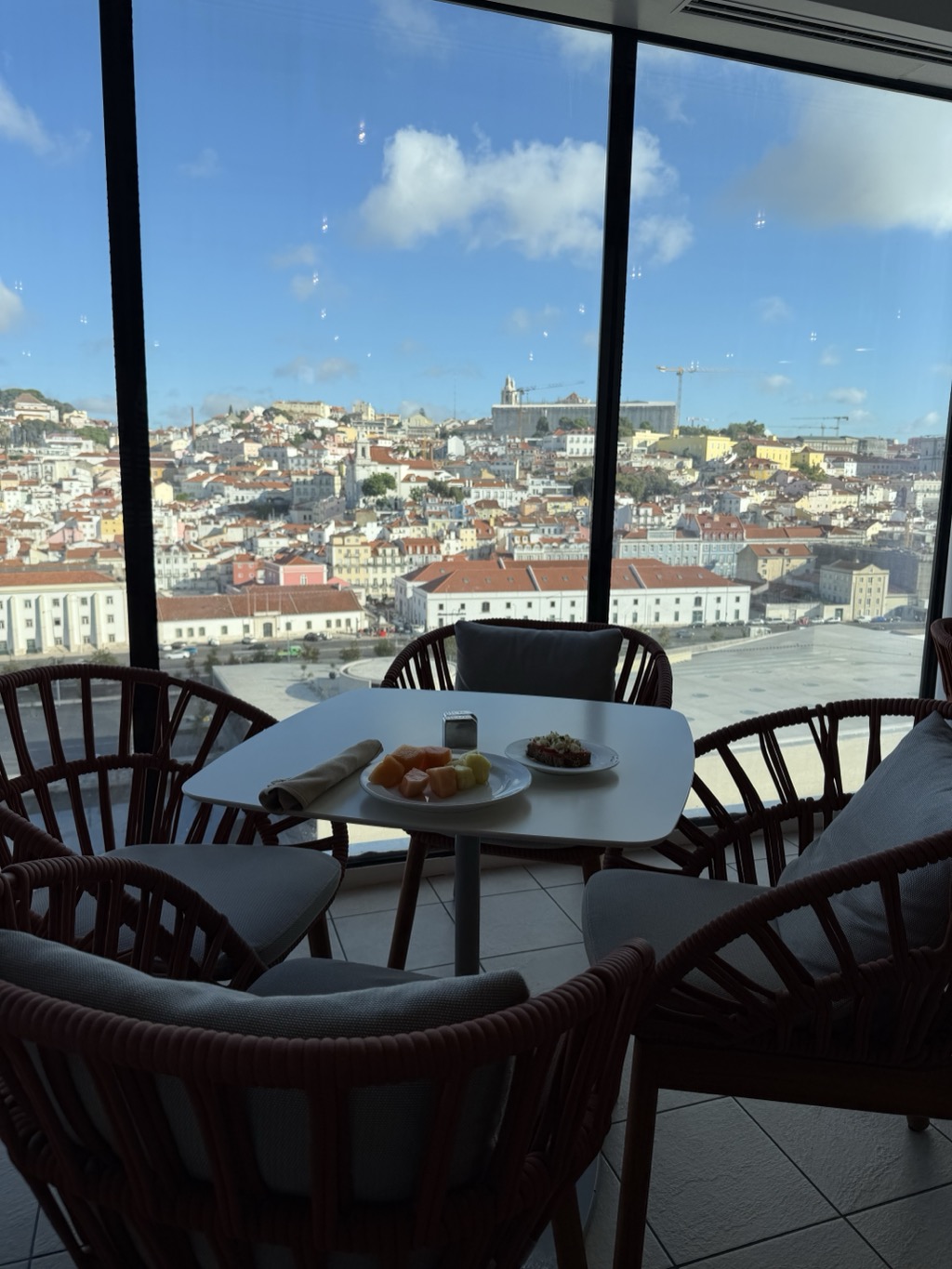 Breakfast at a low white table in a glass-walled shipboard lounge, looking out over Lisbon. On the table sit a white plate of cut melon and pineapple, a second small plate holding a topped toast, a folded napkin, and a metal sugar caddy. Round rope-woven armchairs in dark red with grey cushions surround the table on all sides. Through the floor-to-ceiling windows, the city climbs the hillside in dense tiers of white and pastel buildings under terracotta roofs, with church towers, a large pale monastery-like building, and two yellow construction cranes on the skyline. Blue sky with scattered white cloud.