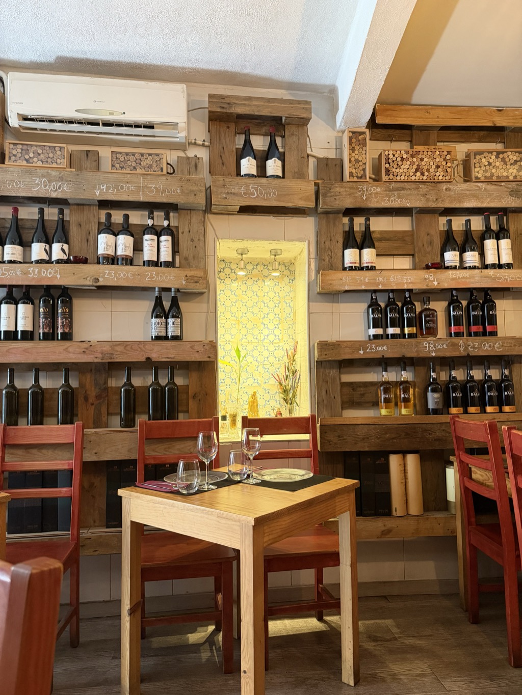 The interior of a small Lisbon restaurant, its walls lined floor to ceiling with shelving built from reclaimed pallet wood and stacked with wine bottles. Prices are chalked directly onto the shelf edges -- 30,00 euros, 42,00, 39,00, 50,00, 23,00, 33,00, 98,00 -- and shallow boxes of used corks sit along the upper shelves. A row of Dalva port bottles with orange labels fills the shelf at the right. In the center, a small niche is lined with blue-and-white patterned tile and holds a plant and a couple of small figurines. A bare pine table in the foreground is set for two with wine glasses, dark placemats, and patterned plates, surrounded by red-painted wooden chairs on a dark wood floor. A white air-conditioning unit is mounted high on the left wall.