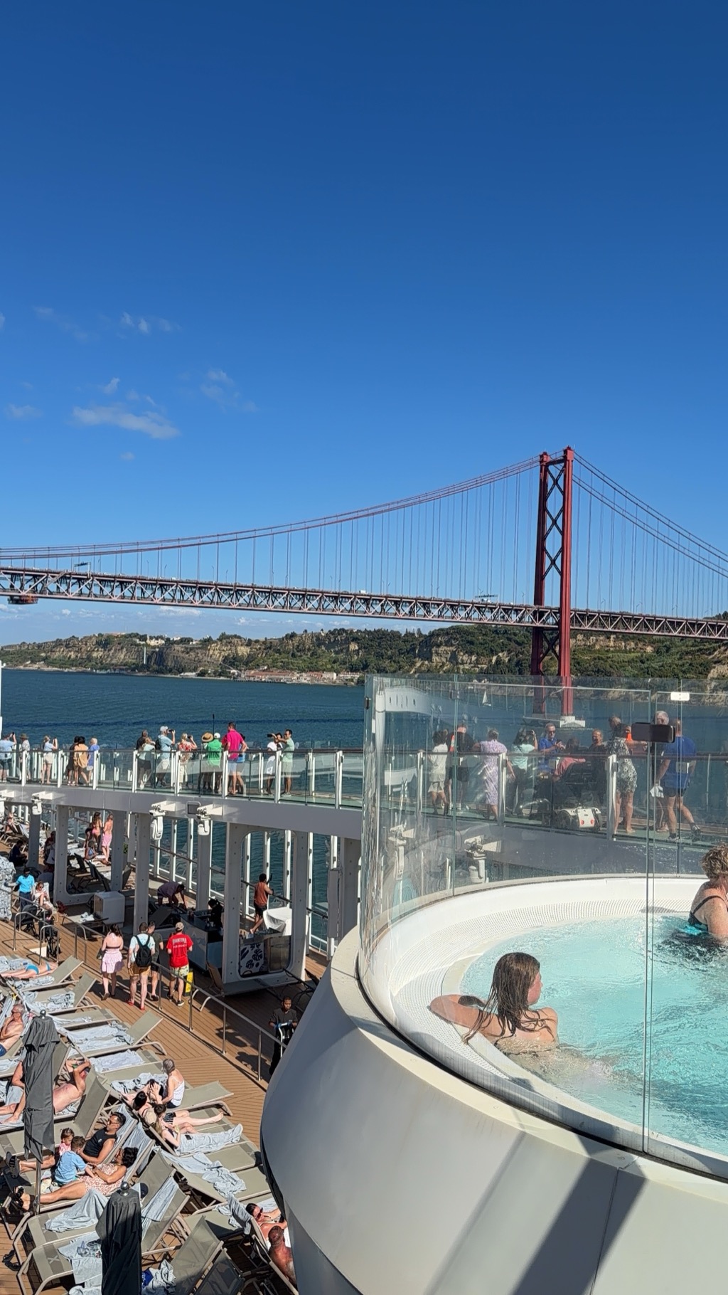 Sailaway on the Tagus, shot from an upper deck of the ship as it passes beneath the 25 de Abril Bridge. The rust-red suspension bridge crosses the upper half of the frame, its tower and cables rising against a deep blue sky, with the wooded green hillside of the south bank and the white buildings of Almada below it. In the foreground at right, passengers sit in a circular glass-walled hot tub cantilevered over the side; along the railings above and to the left, dozens of people stand watching and photographing the bridge. The teak pool deck below is filled with rows of lounge chairs, most occupied by sunbathers, and crew and passengers move along the deck.